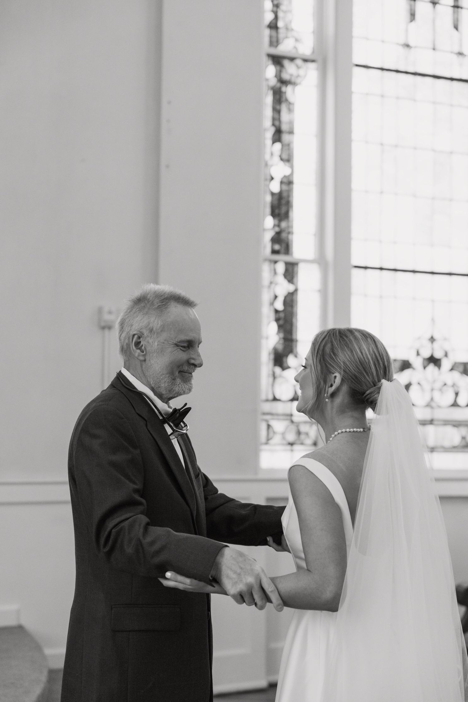 Black and white photo of a smiling groom holding hands with a bride in a veil at a church wedding ceremony.