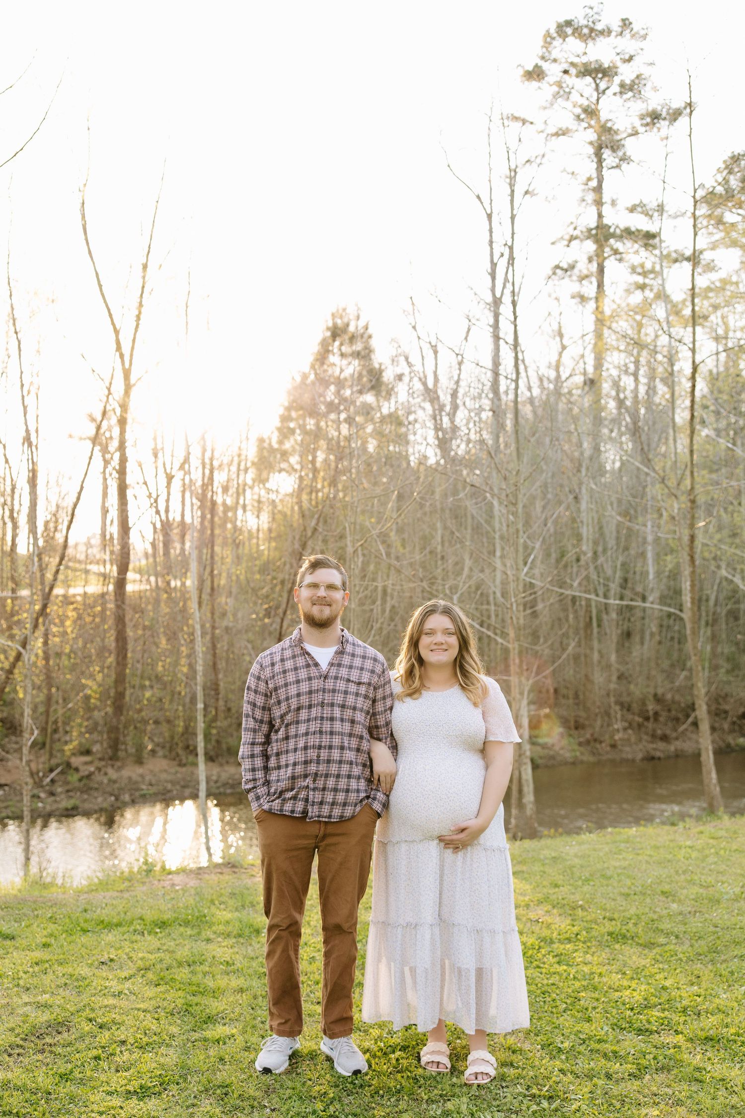 Pregnant couple posing outdoors at golden hour near a pond surrounded by trees, woman in white dress cradling baby bump.
