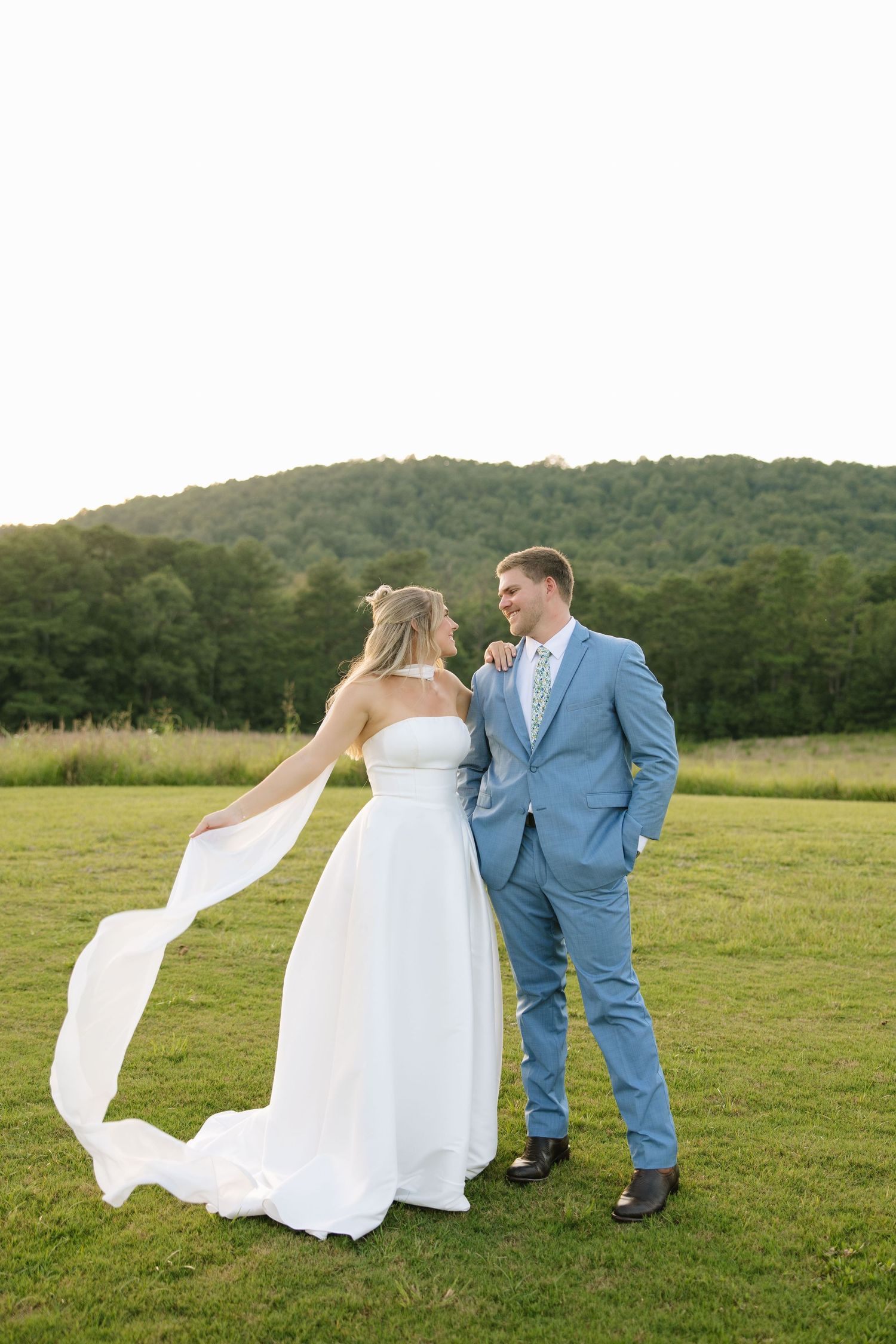 Bride in flowing white gown and groom in blue suit share a romantic moment in a lush green field with wooded hills behind.