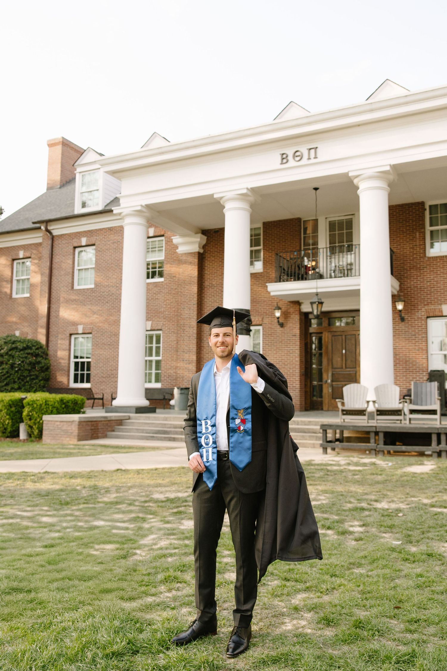 Graduate in cap and gown poses proudly in front of Beta Theta Pi fraternity house on college campus.