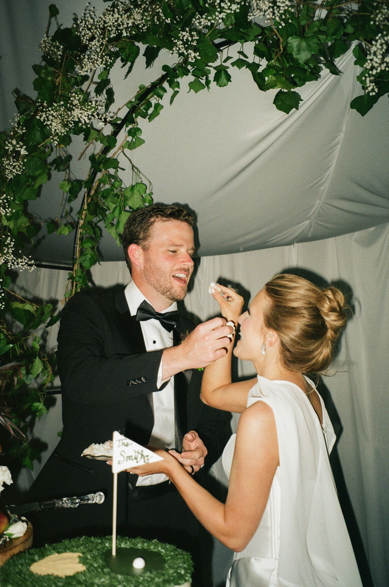 Bride and groom sharing cake at wedding reception under floral greenery arch, groom in tuxedo, bride in white gown.