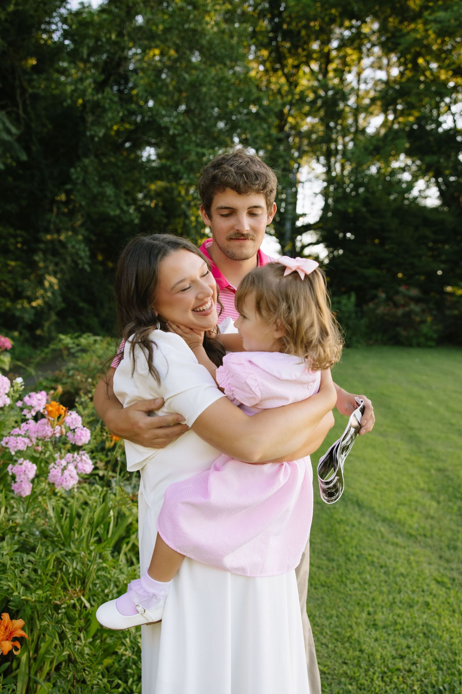 Happy young family with mother, father, and toddler daughter in pink dress posing outdoors near blooming flowers.