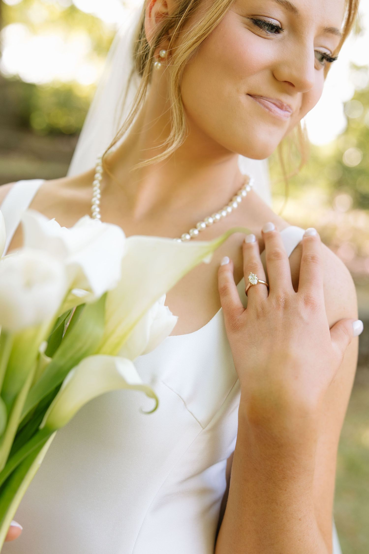 Bride in white gown with pearl necklace and ring holding white calla lily bouquet outdoors.