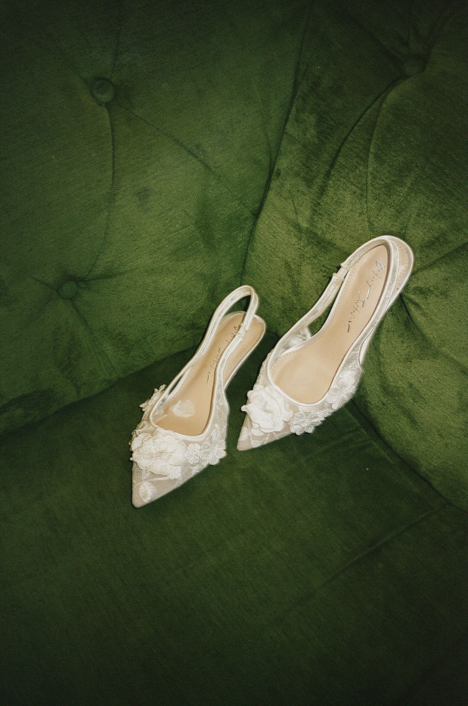 Elegant white lace slingback heels with floral embellishments resting on a tufted green velvet sofa.