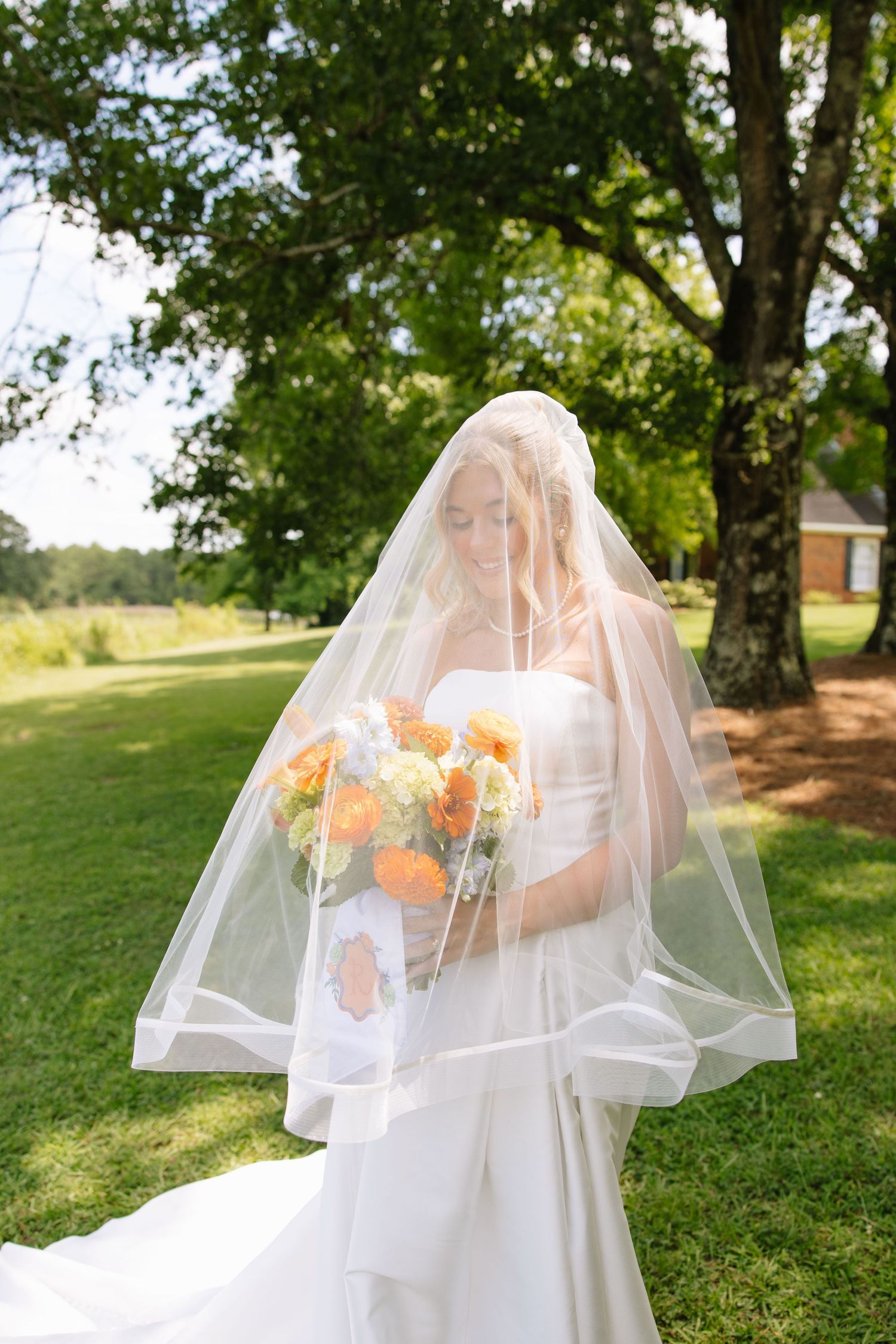 Bride in white gown and veil holding orange and yellow bouquet outdoors on green lawn with trees.