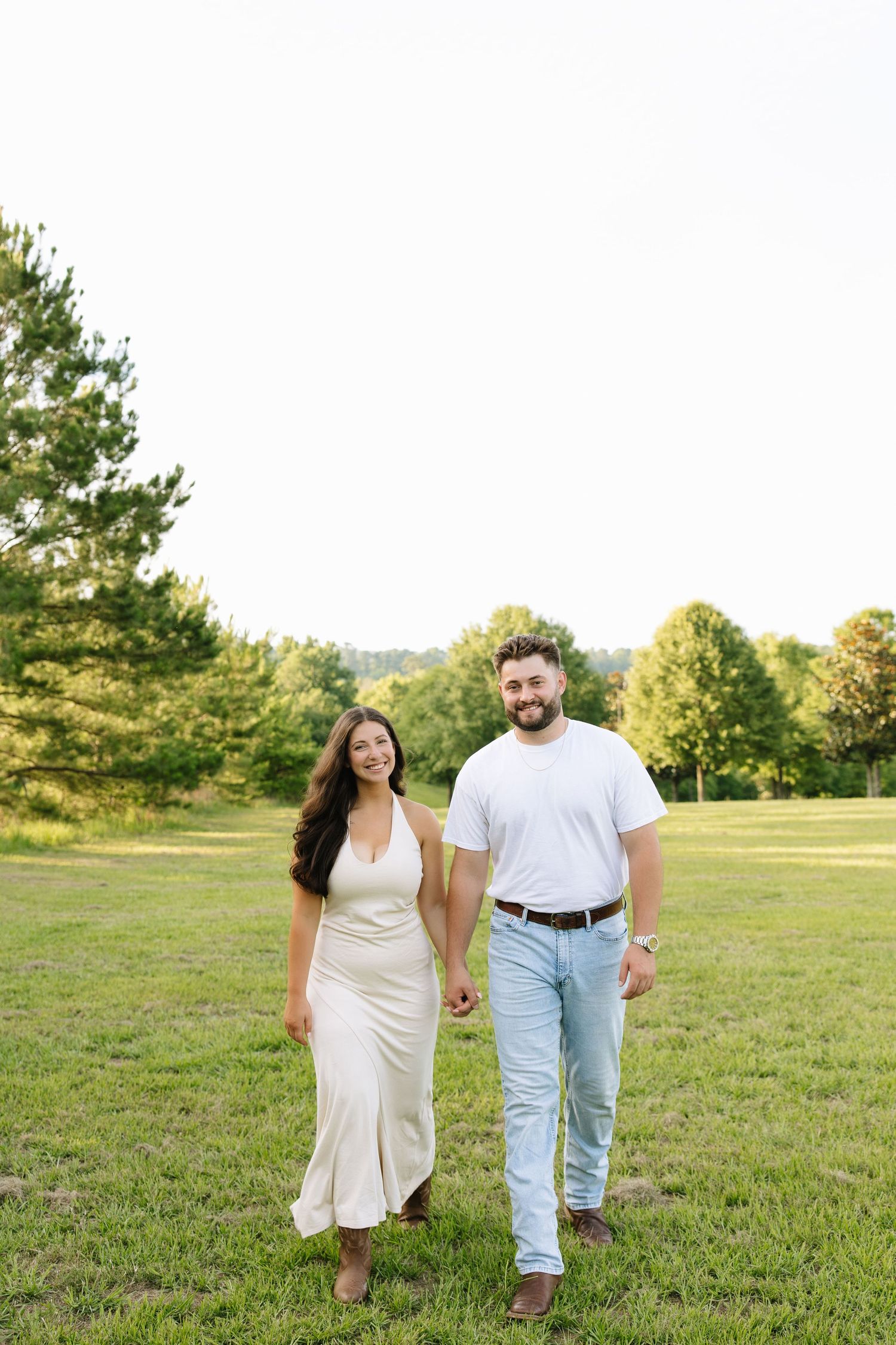 A couple holds hands while walking through a lush green field, woman in cream dress, man in white tee and light jeans.
