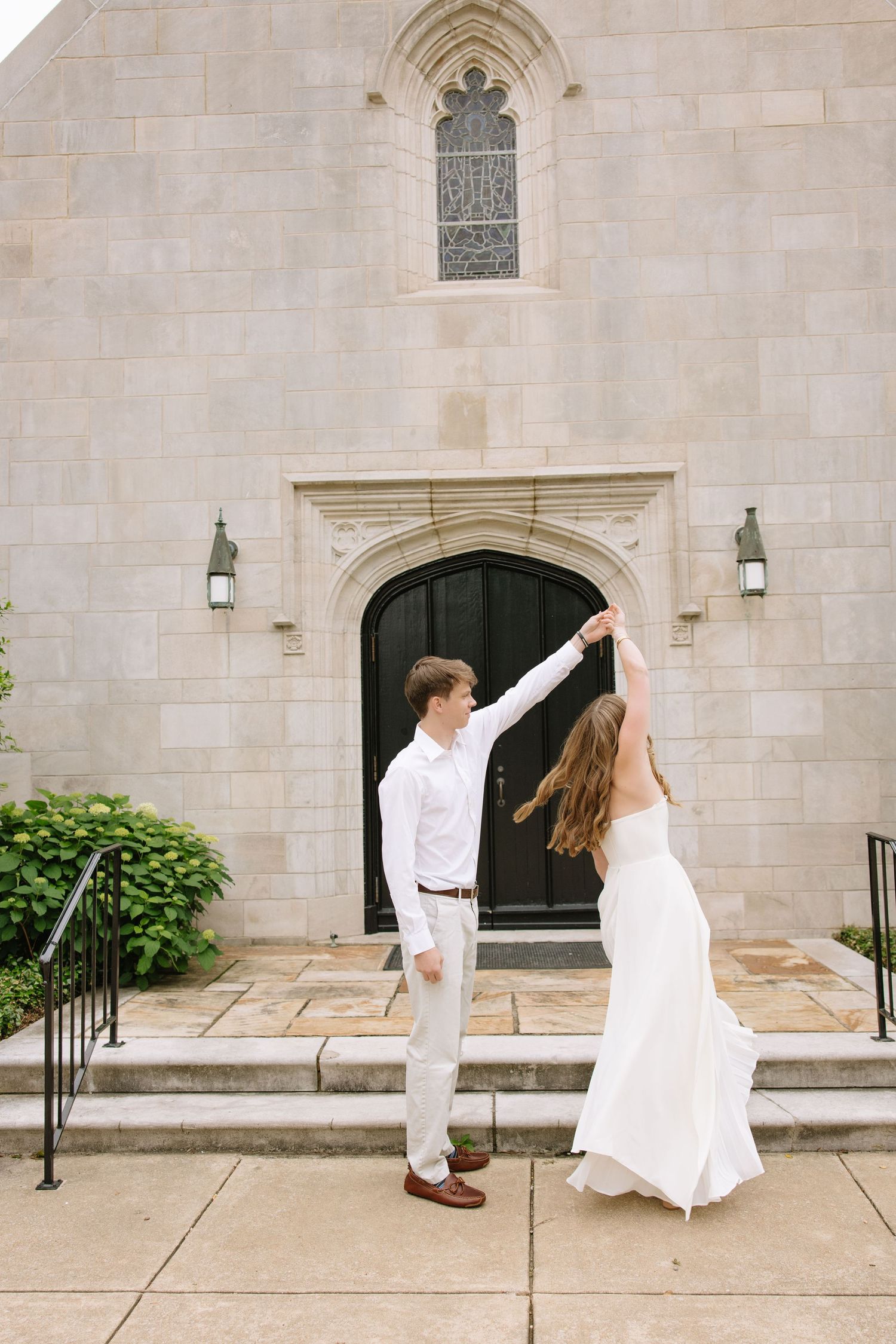 Bride and groom dancing outside a stone church, bride in white gown, groom in white shirt and gray pants.
