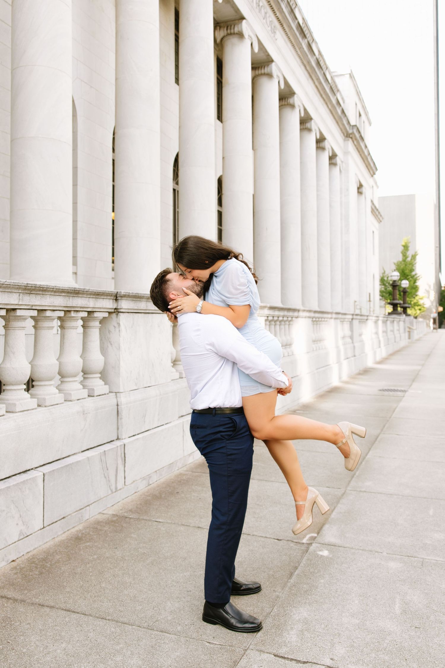 Couple sharing a romantic kiss, man lifting woman in front of elegant white stone columns outdoors.