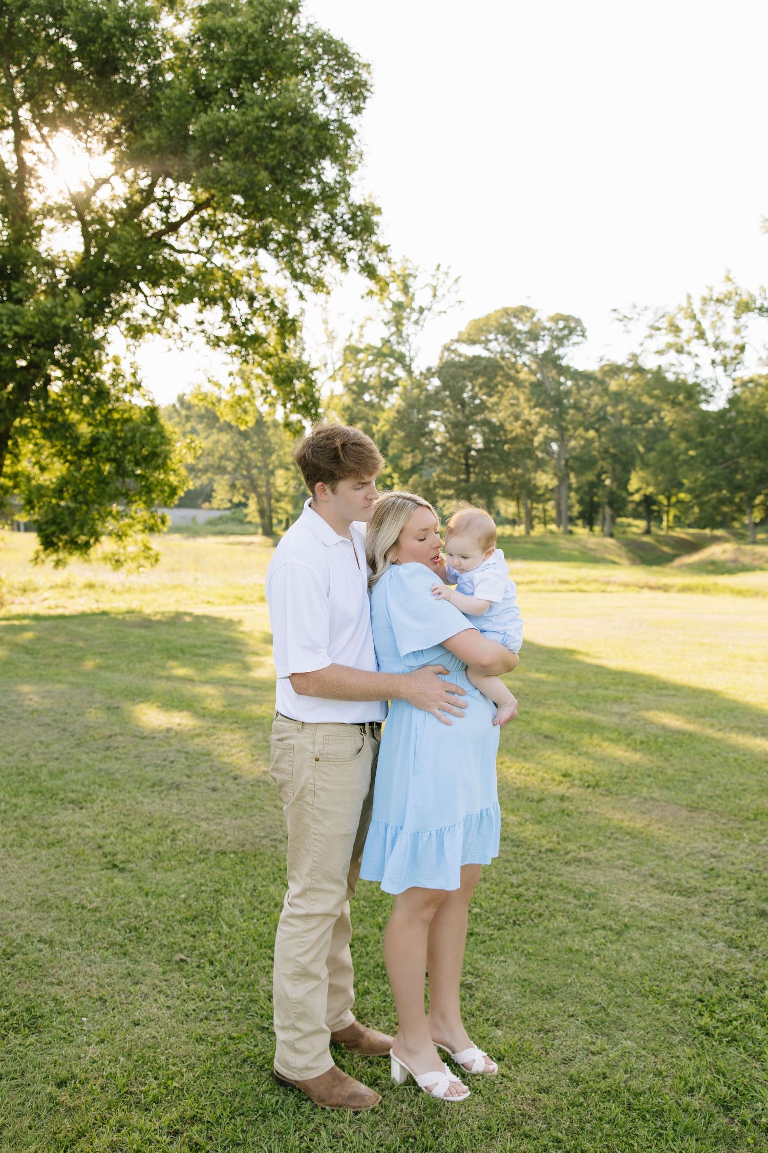 Couple sharing tender moment with baby in sunny park, woman in blue dress, man in white shirt and khakis.