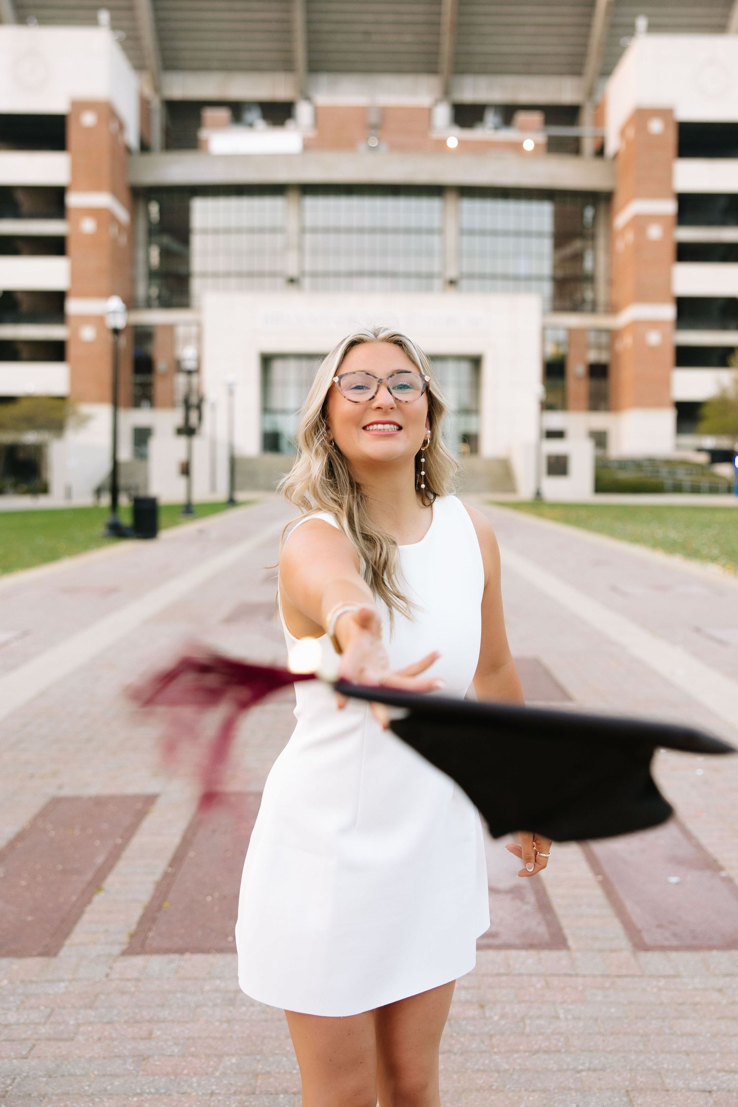 Smiling graduate in white dress tossing cap outside university stadium.