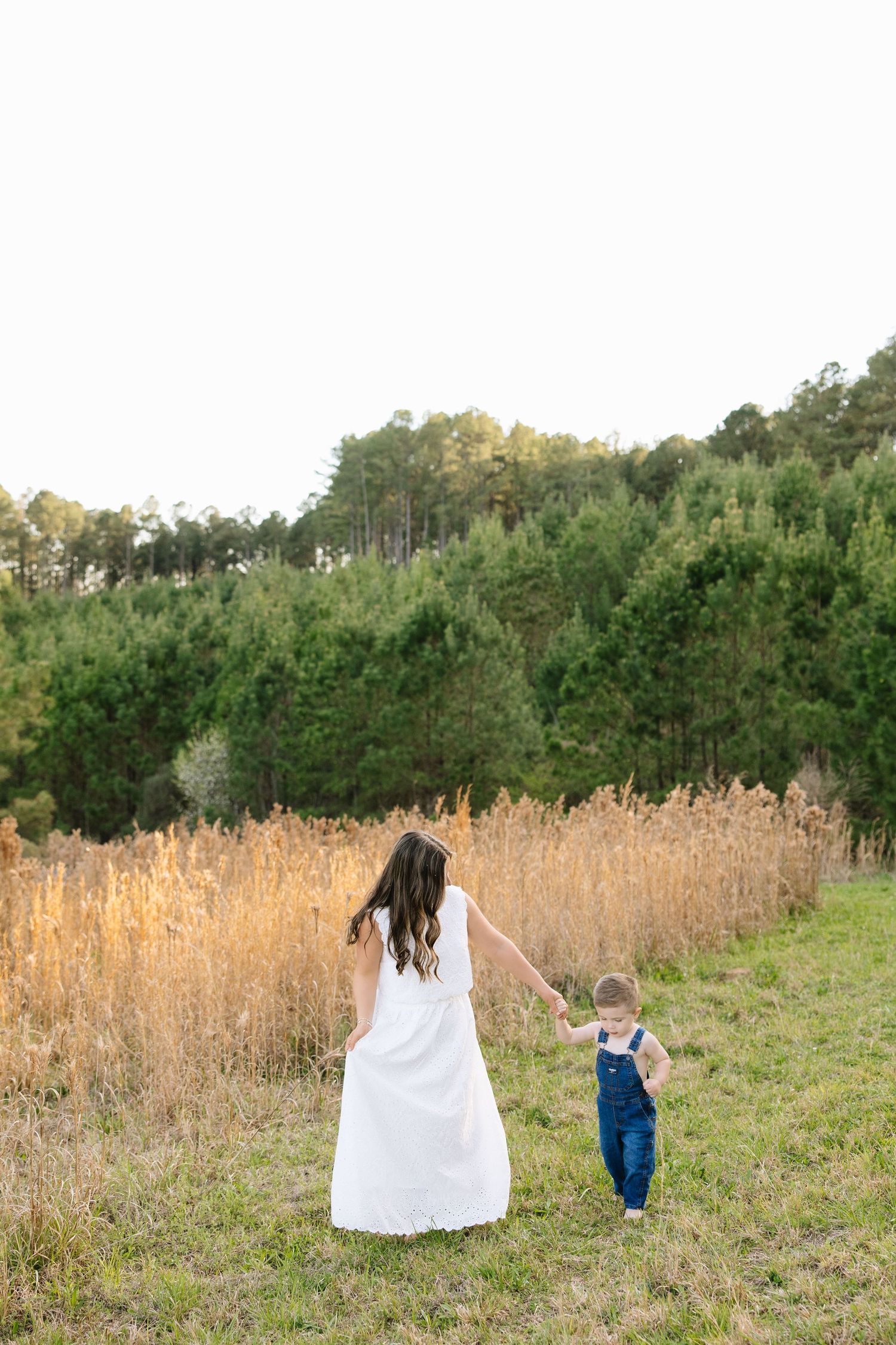 A mother in a white dress holds hands with her toddler in denim overalls walking through a sunny field.