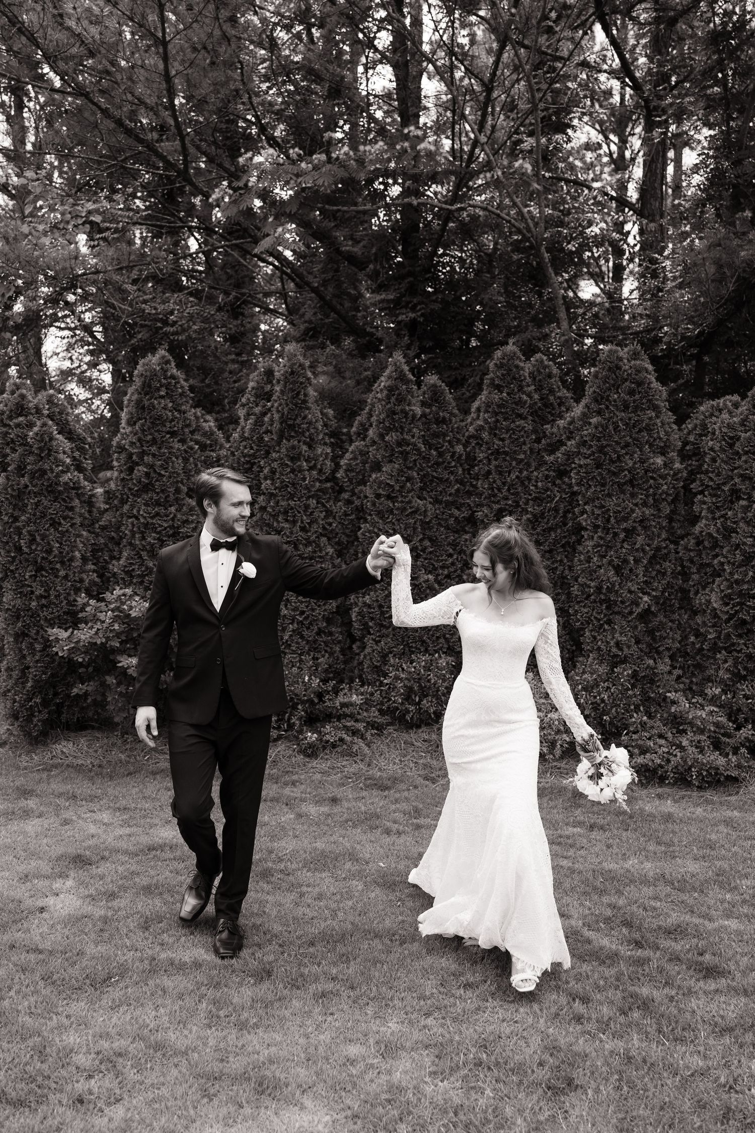 A black and white wedding photo showing a couple walking hand in hand on grass with evergreen trees in the background.