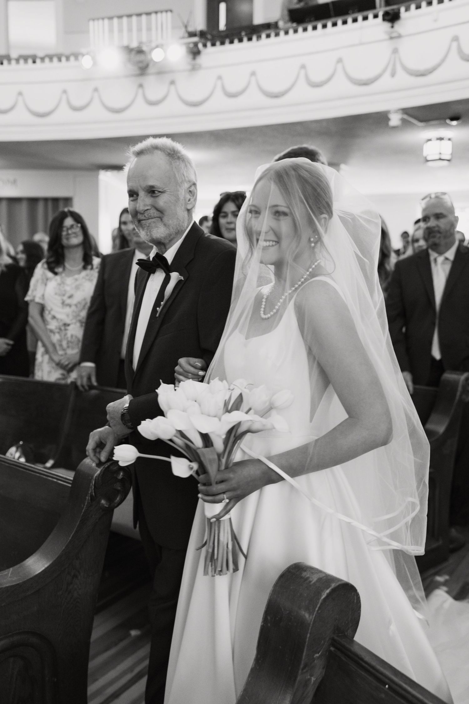 Black and white photo of smiling bride walking down church aisle with bouquet, escorted by older gentleman.