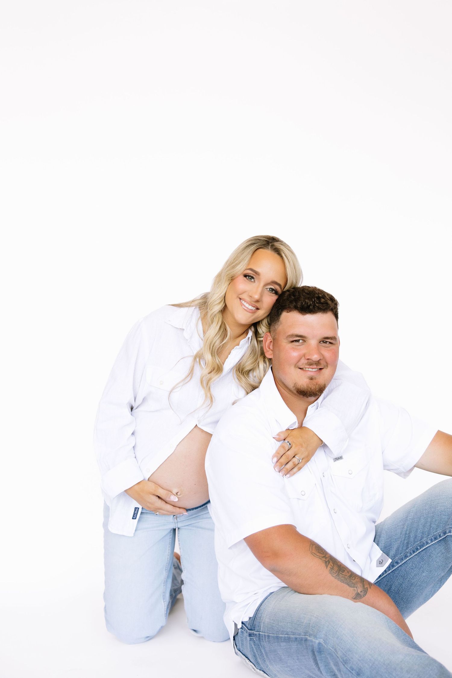 Pregnant couple in white shirts and jeans posing together on white background, woman kneeling behind man smiling.
