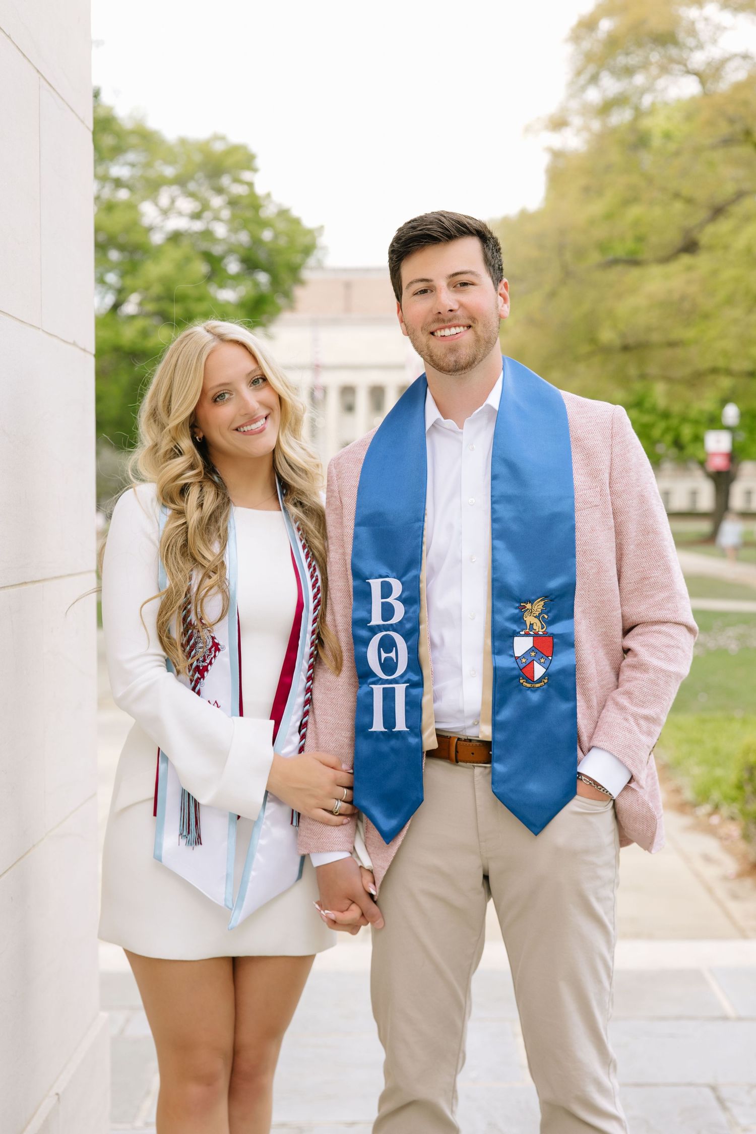 Two smiling college graduates pose together wearing graduation stoles, the man wearing a Beta Theta Pi fraternity sash.