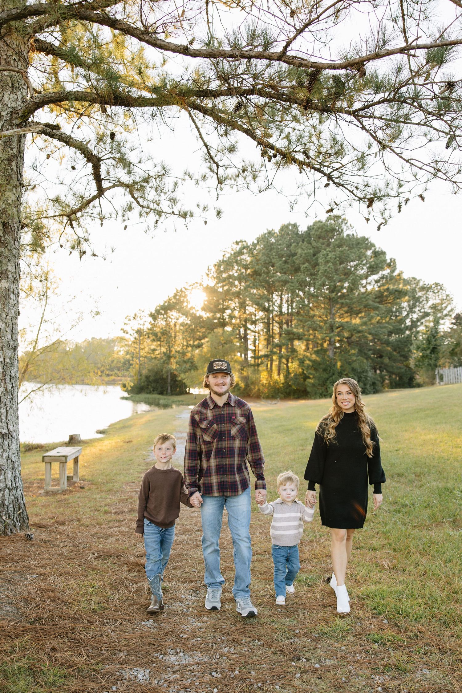 Family of four posing on a scenic path by a lake at golden hour, surrounded by tall pine trees in autumn.