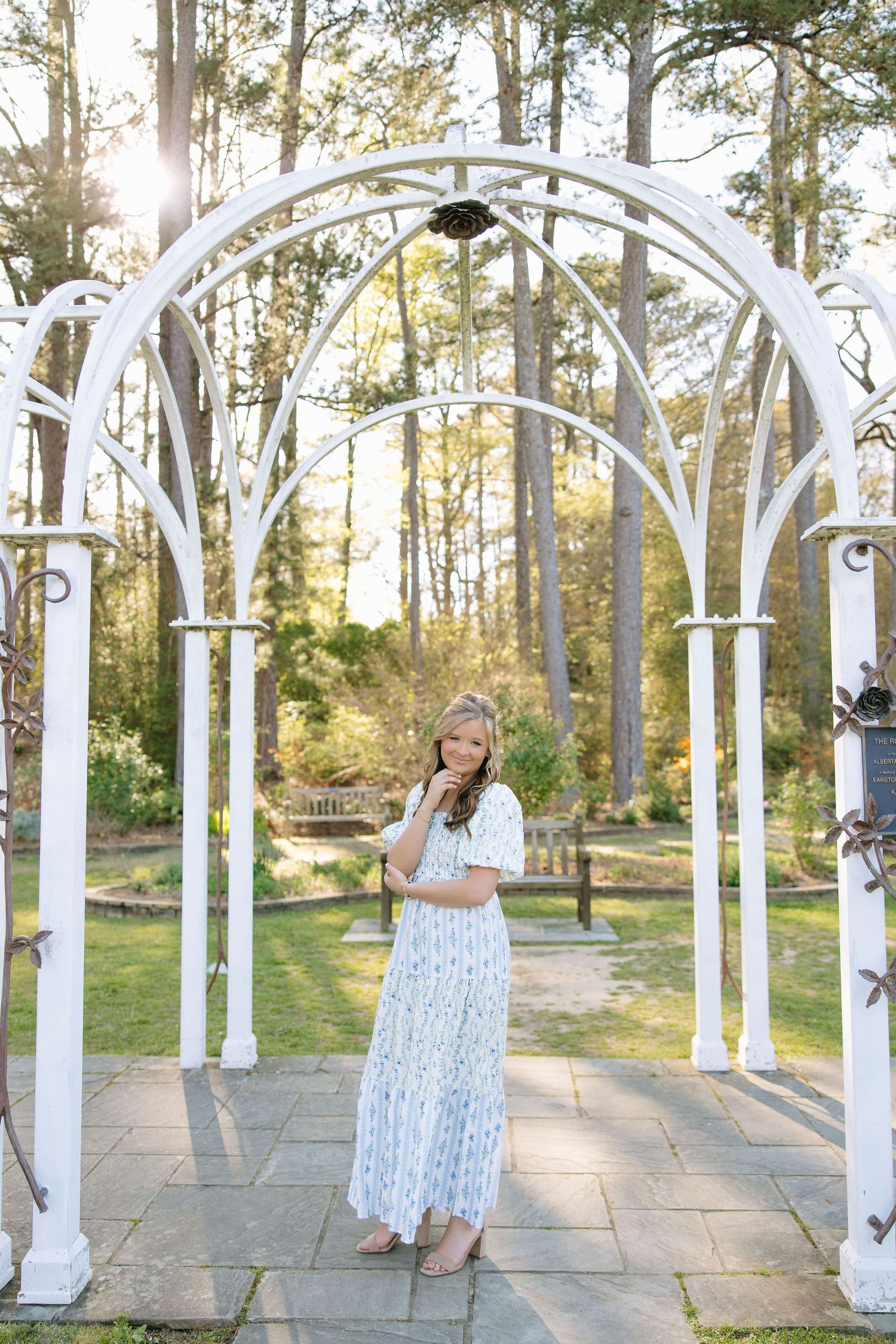 Young woman in a blue floral maxi dress standing under a white garden arch surrounded by lush trees in golden sunlight.