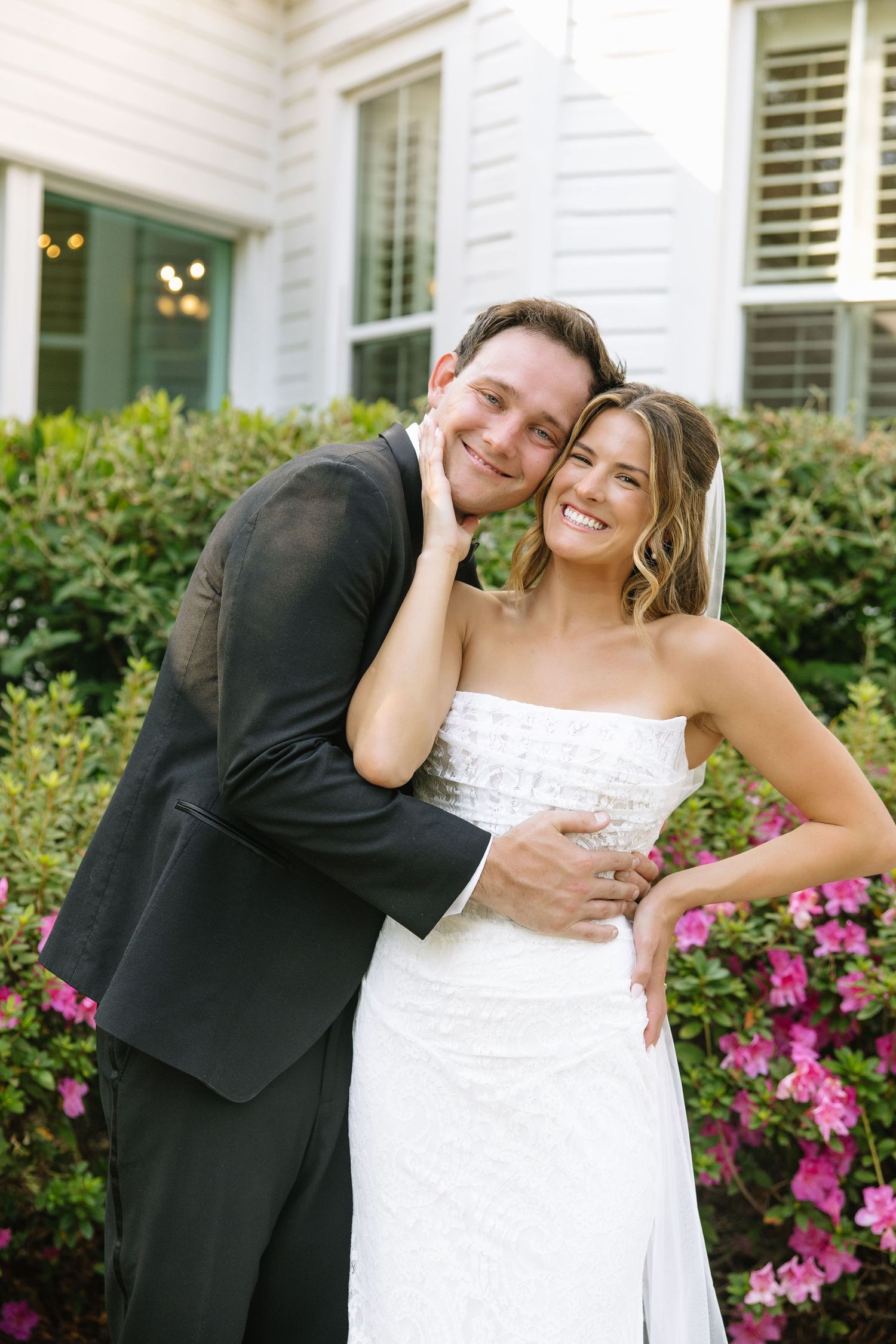 Playful wedding portraits taken outside a white house with blooming pink flowers.