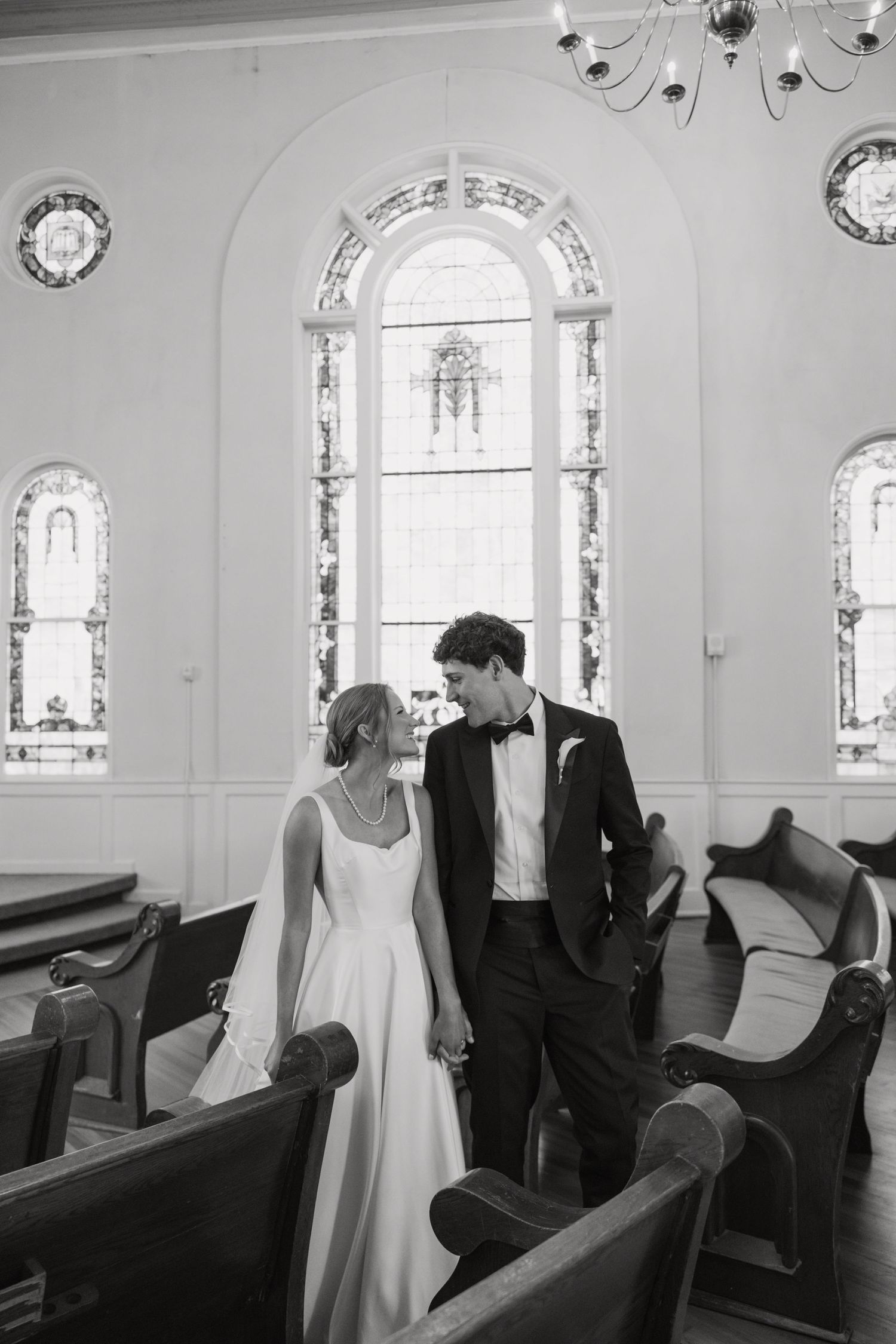 Black and white photo of a bride and groom holding hands in a church aisle with stained glass windows.