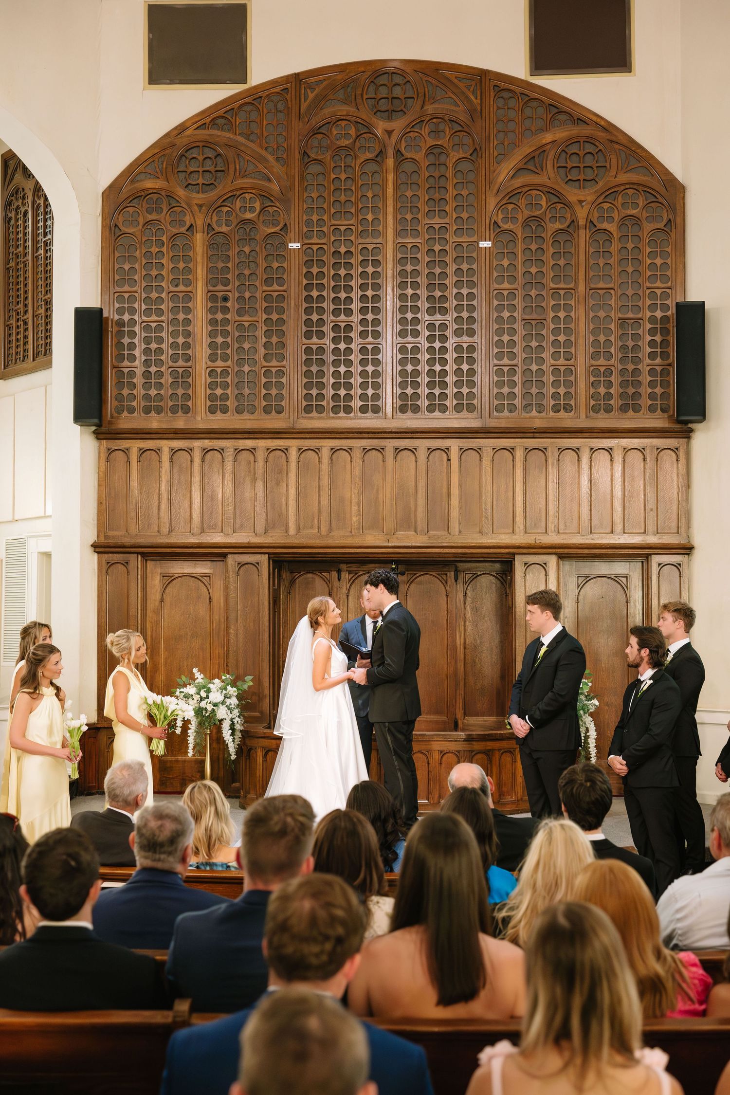 Bride and groom exchange vows at church altar with wedding party beside ornate wooden backdrop.
