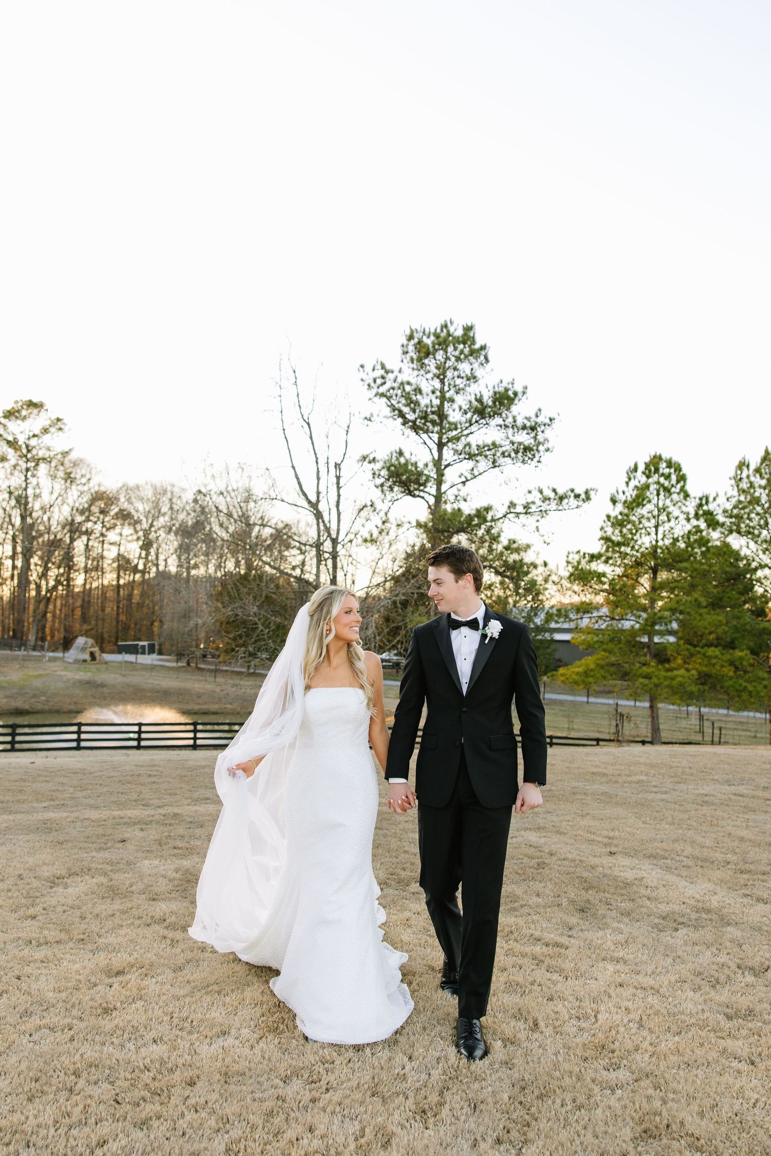 Newlyweds walking together on rustic grounds with pine trees in background.