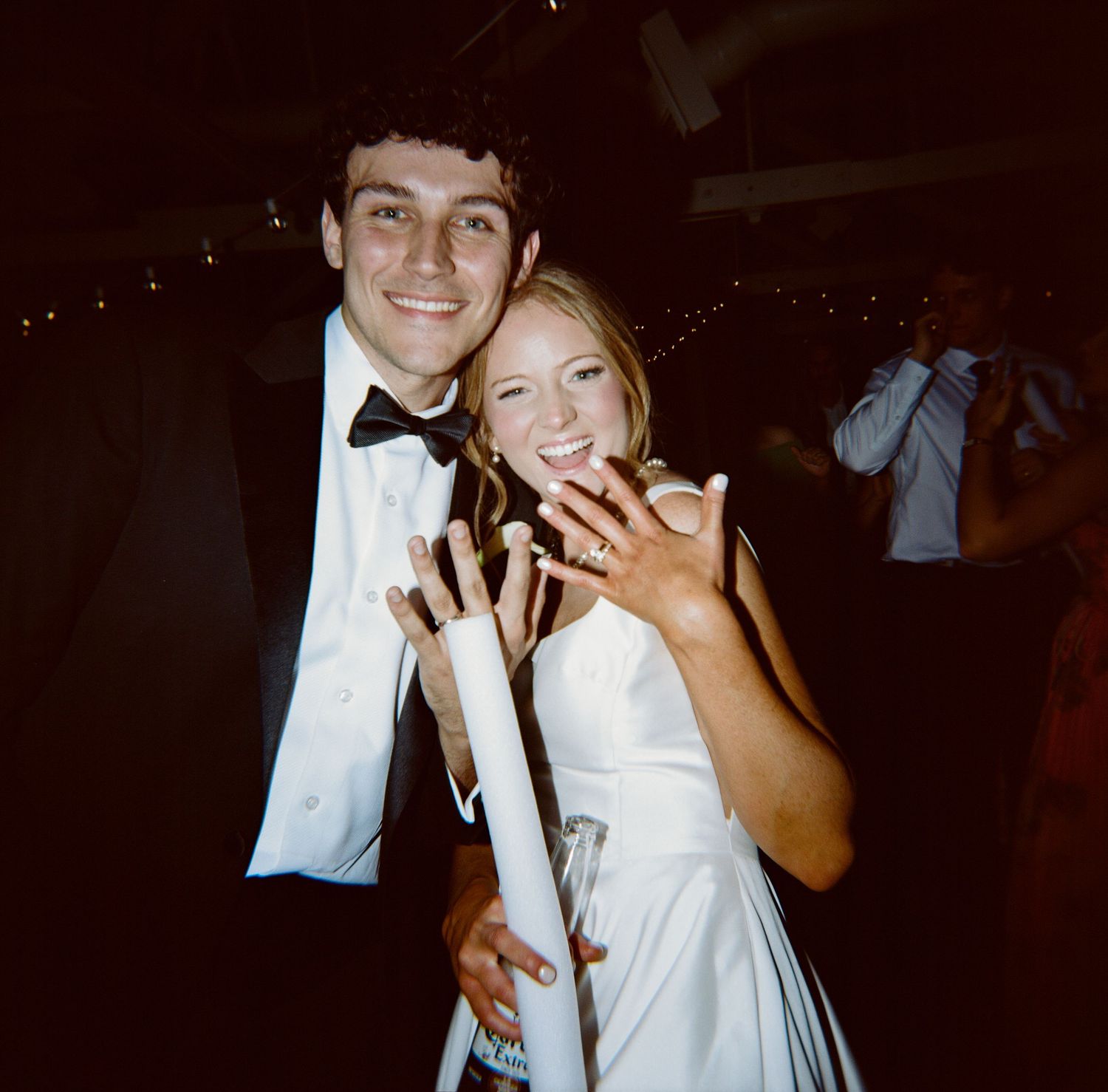 Happy bride and groom showing off wedding rings at a lively indoor wedding reception.