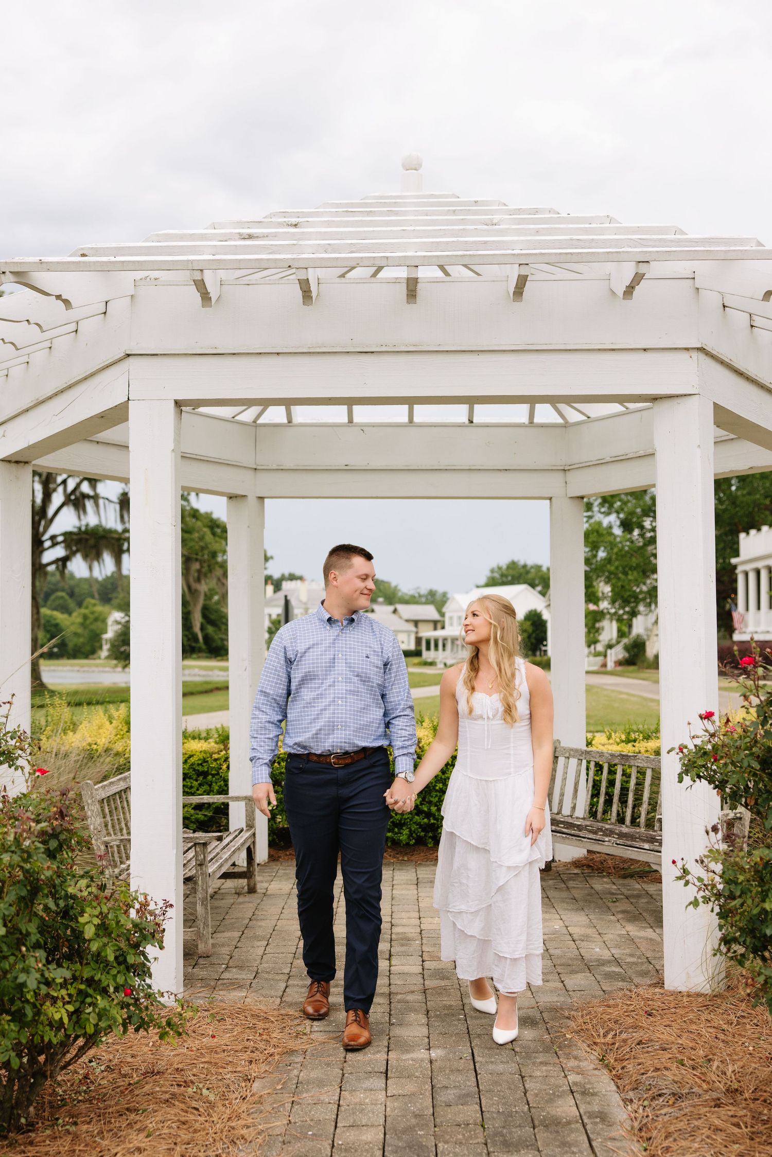 Couple holding hands walking under white pergola gazebo, woman in white dress and man in blue shirt, outdoor garden setting.