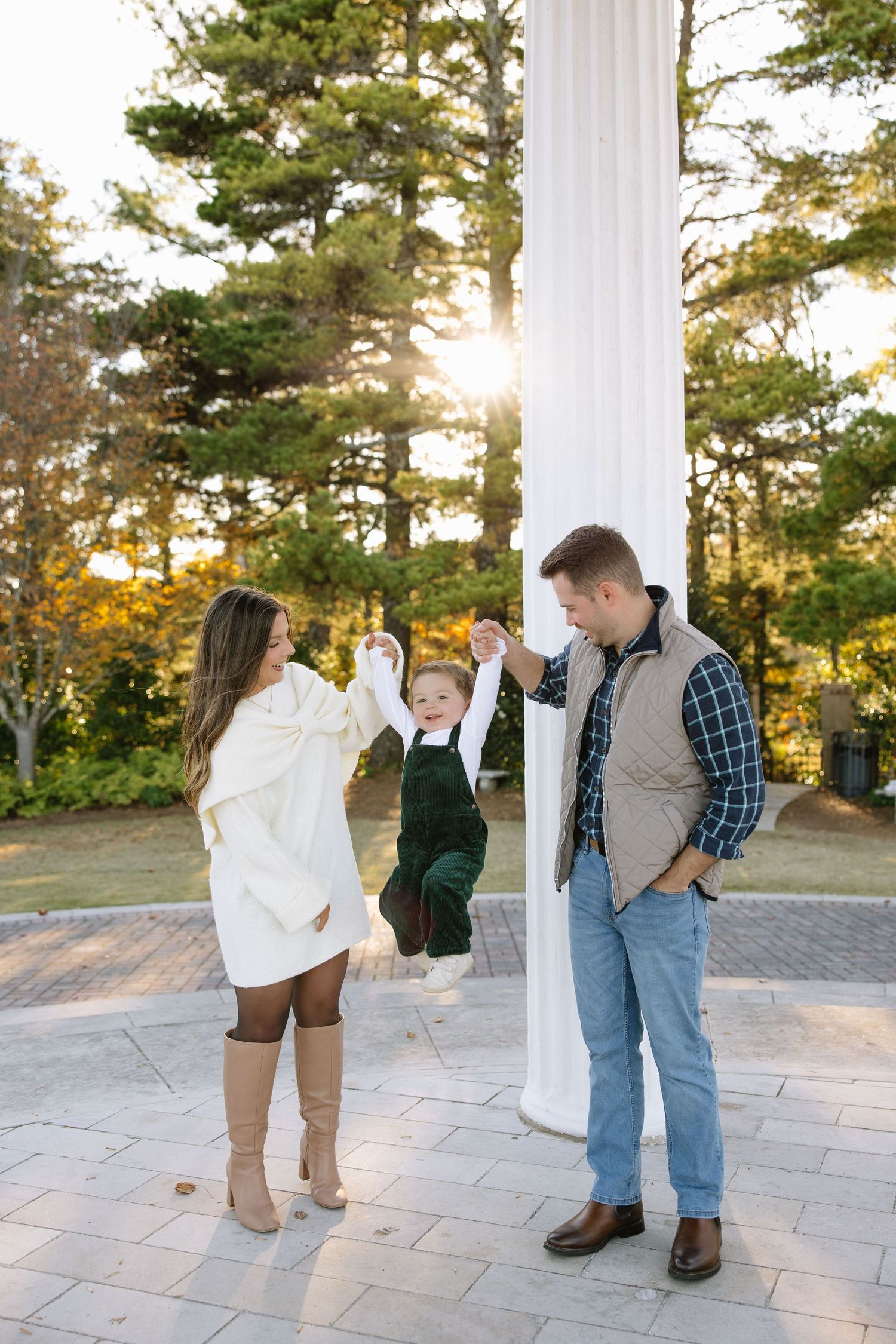 Family of three swinging toddler between parents outdoors near white column with sunlit autumn trees in background.