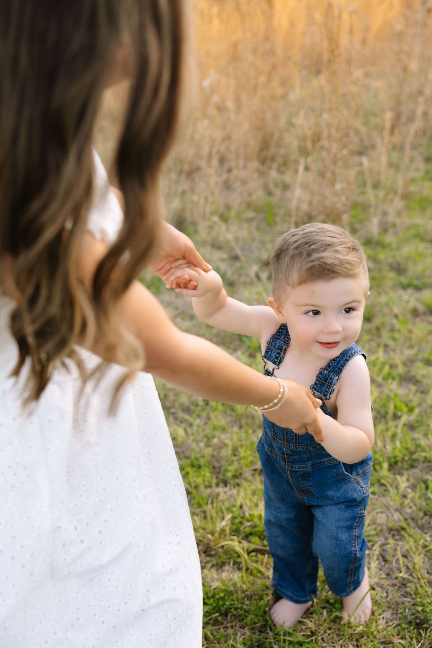 A toddler boy in denim overalls holds his mother's hand in a sunny field, smiling warmly.