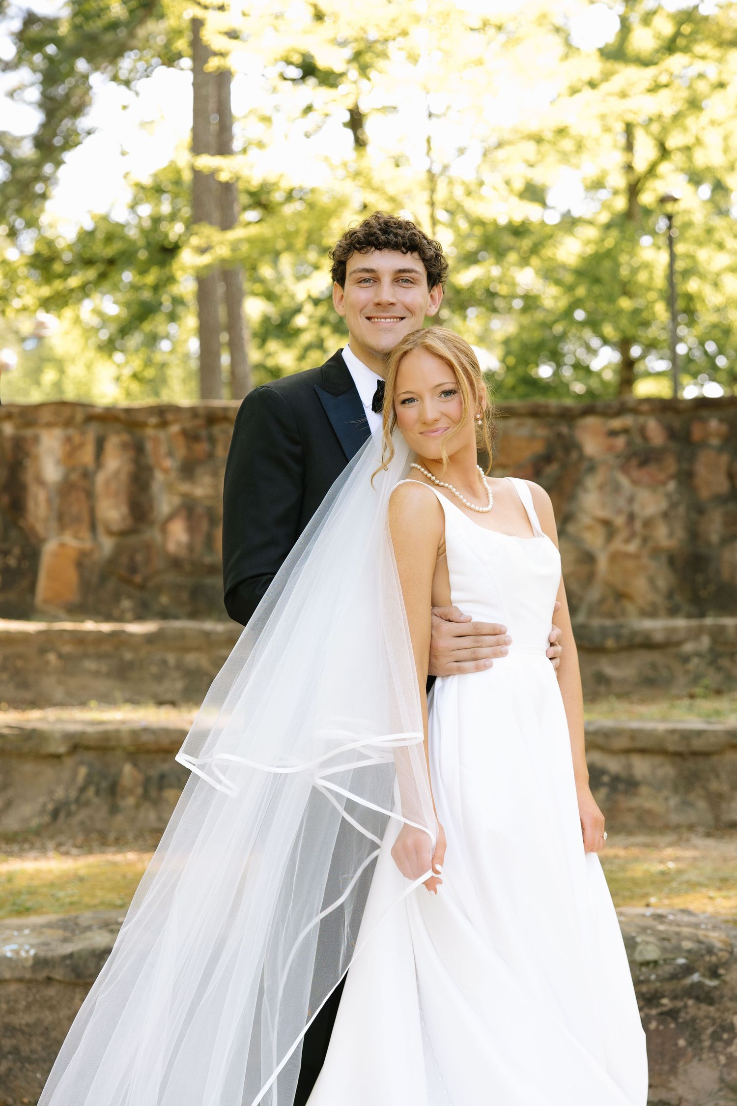 A smiling bride in a white gown and veil poses with her groom in a black tuxedo outdoors among lush green trees.