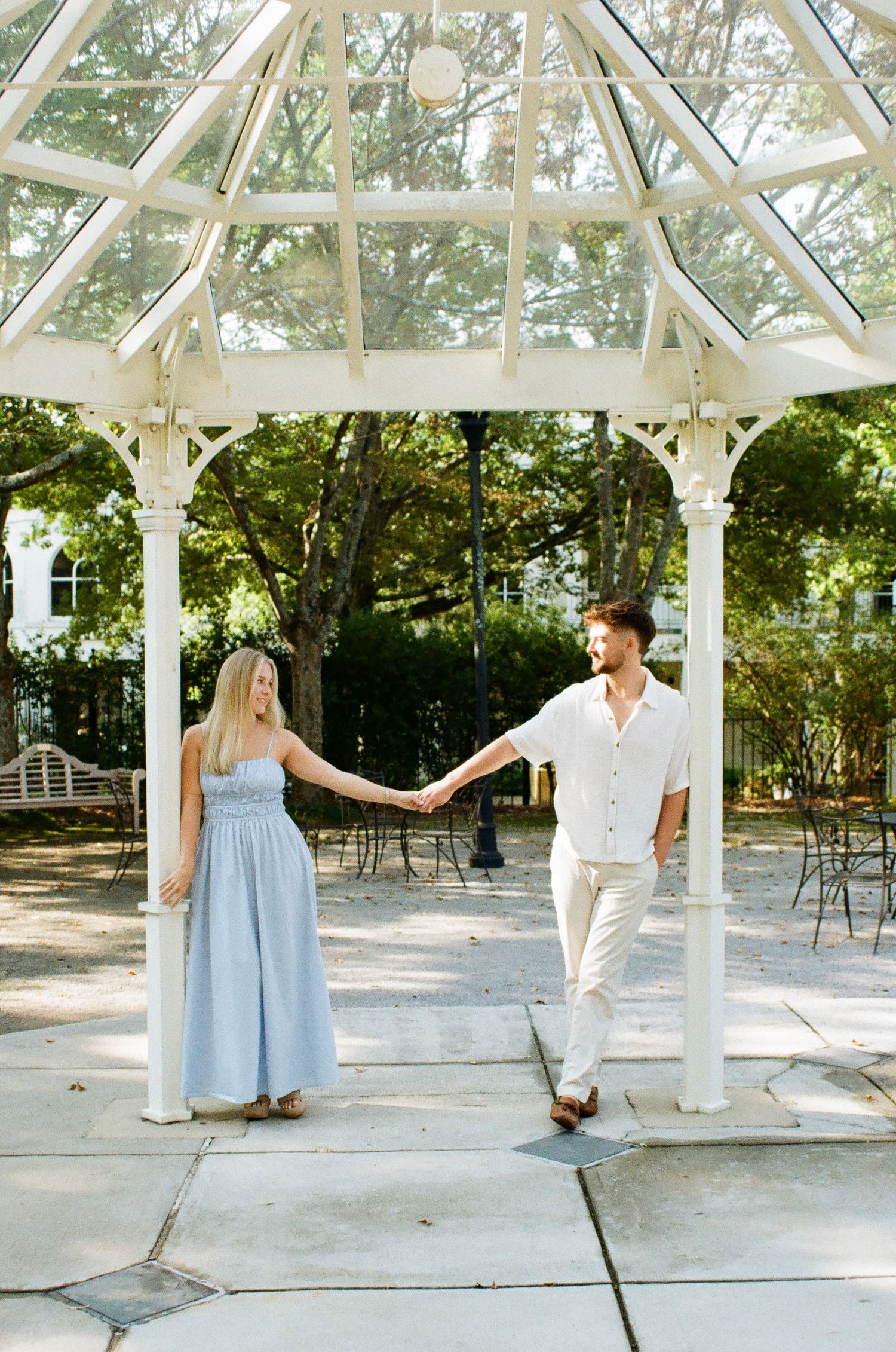 Couple holding hands under a white gazebo with glass roof, woman in blue dress, man in white outfit, outdoor garden setting.