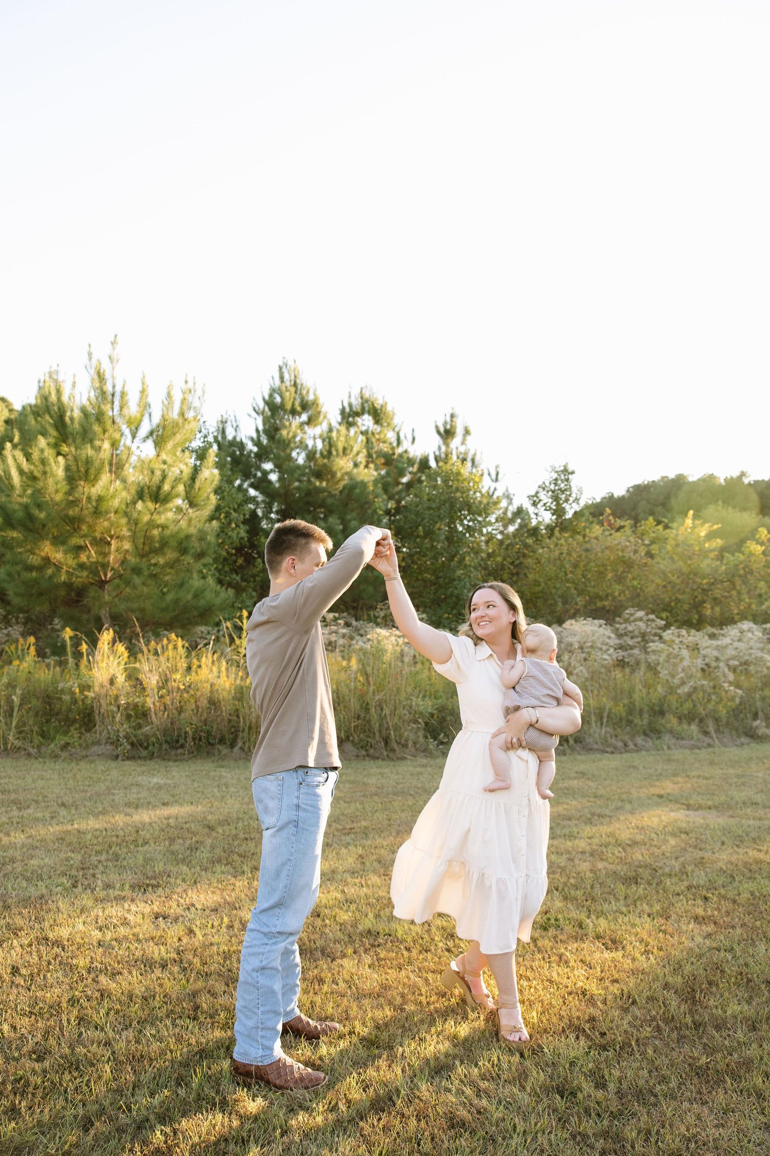 Happy young family dancing in a sunny meadow, mother in white dress holding baby while father spins her hand.