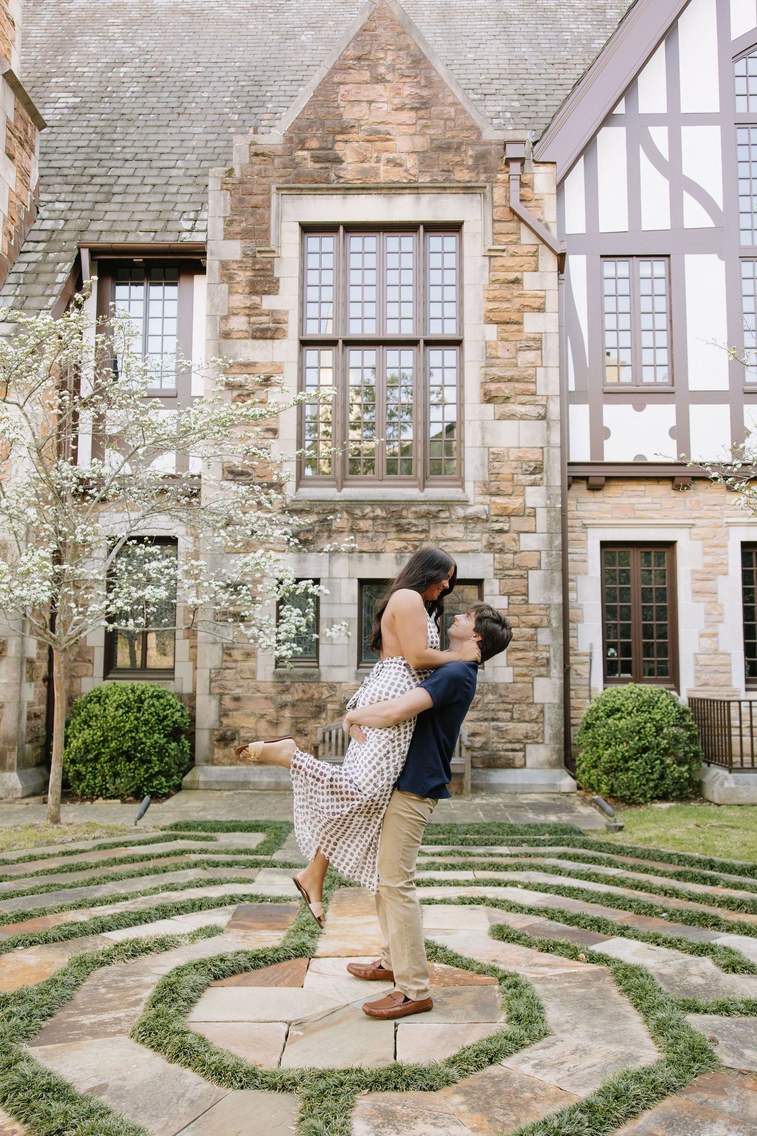 Couple sharing a romantic lift embrace in a Tudor-style garden courtyard with blooming trees and manicured hedges.