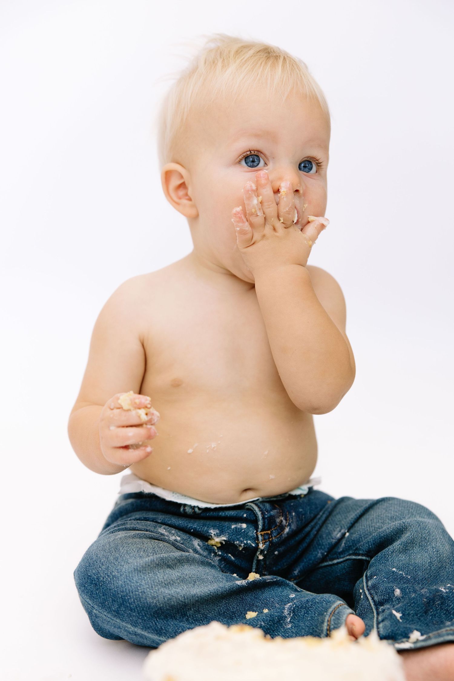Messy toddler with blue eyes eating cake with hands, wearing jeans, sitting against white background.