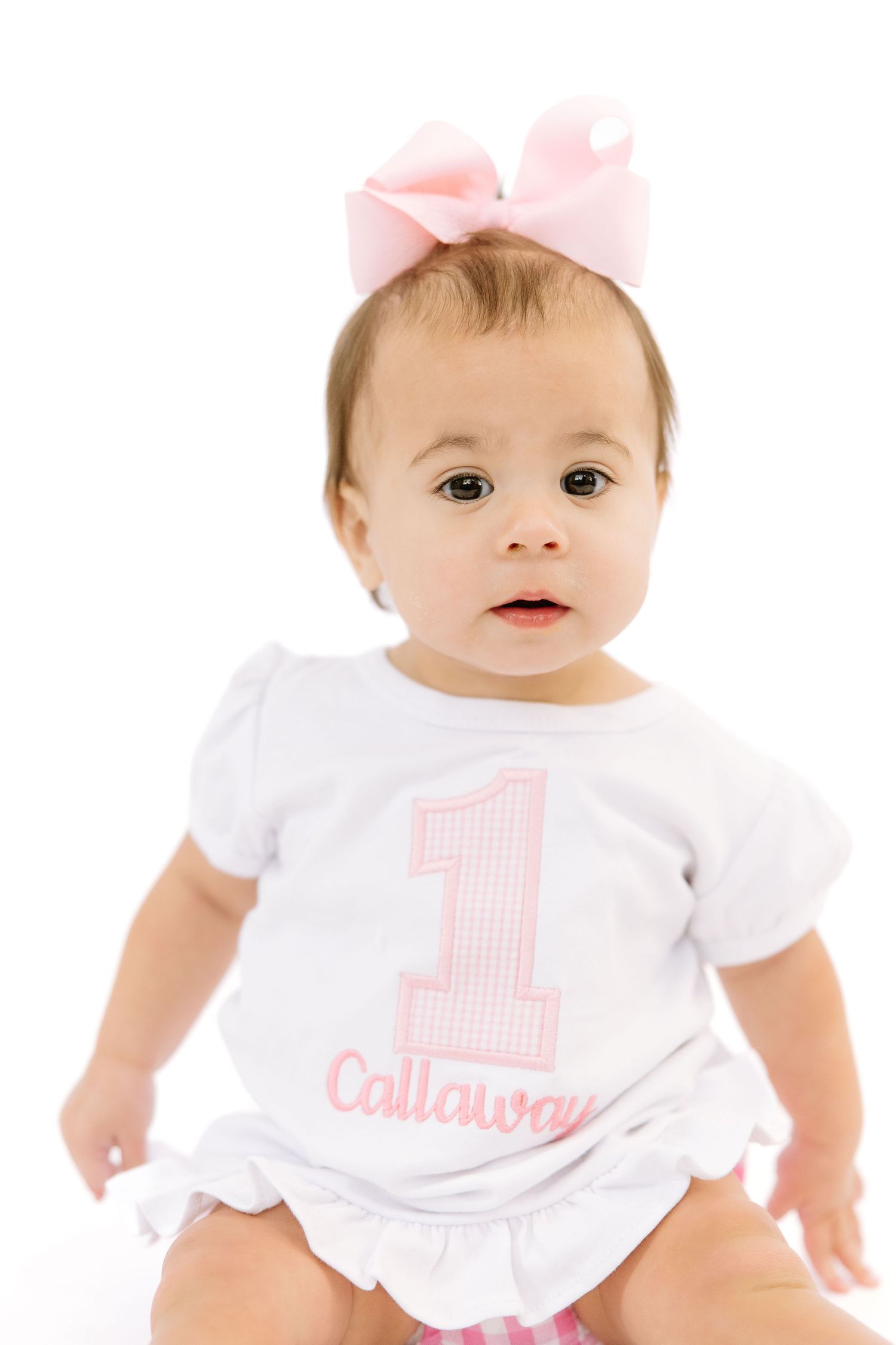 Baby girl in white birthday shirt with pink number 1 and name Callaway, wearing pink bow headband.