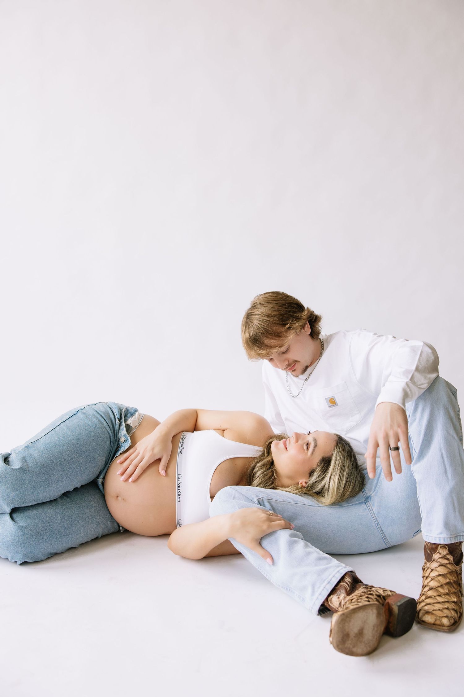 Pregnant woman lying on floor with partner during maternity photo shoot in white studio setting.