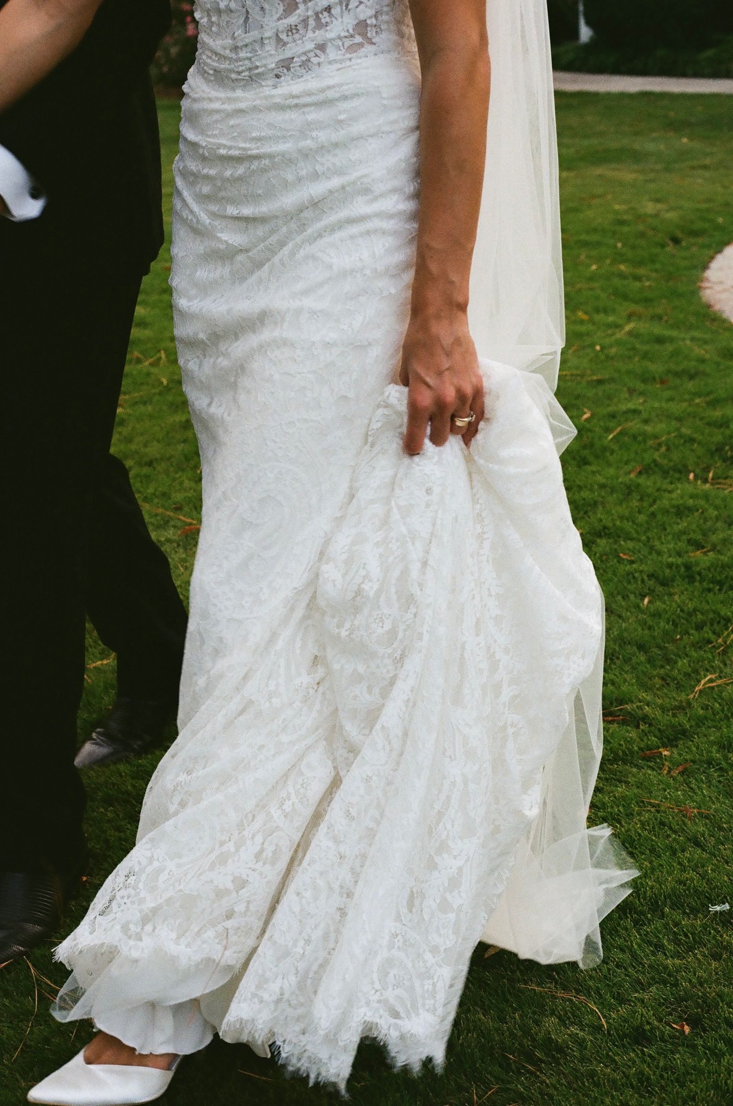 Close-up view of a bride's white lace wedding dress hem and shoes on grass during an outdoor ceremony.