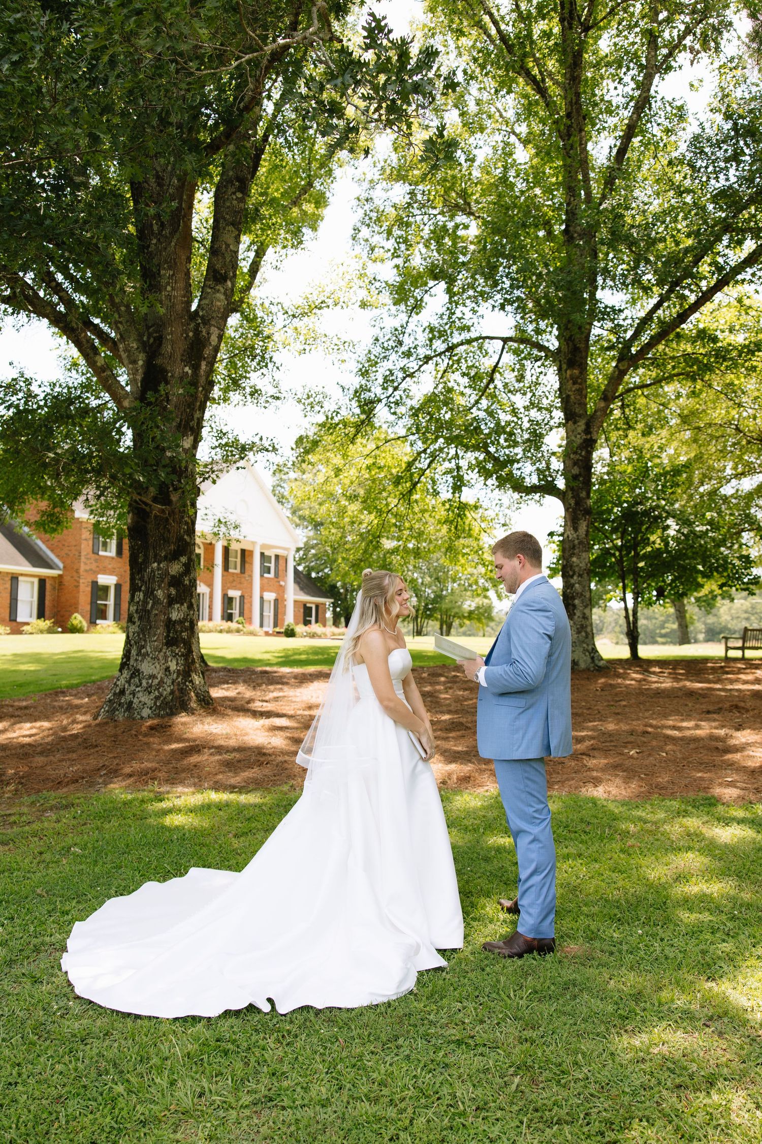 Bride in white gown and groom in blue suit exchange vows outdoors under large trees on green lawn.