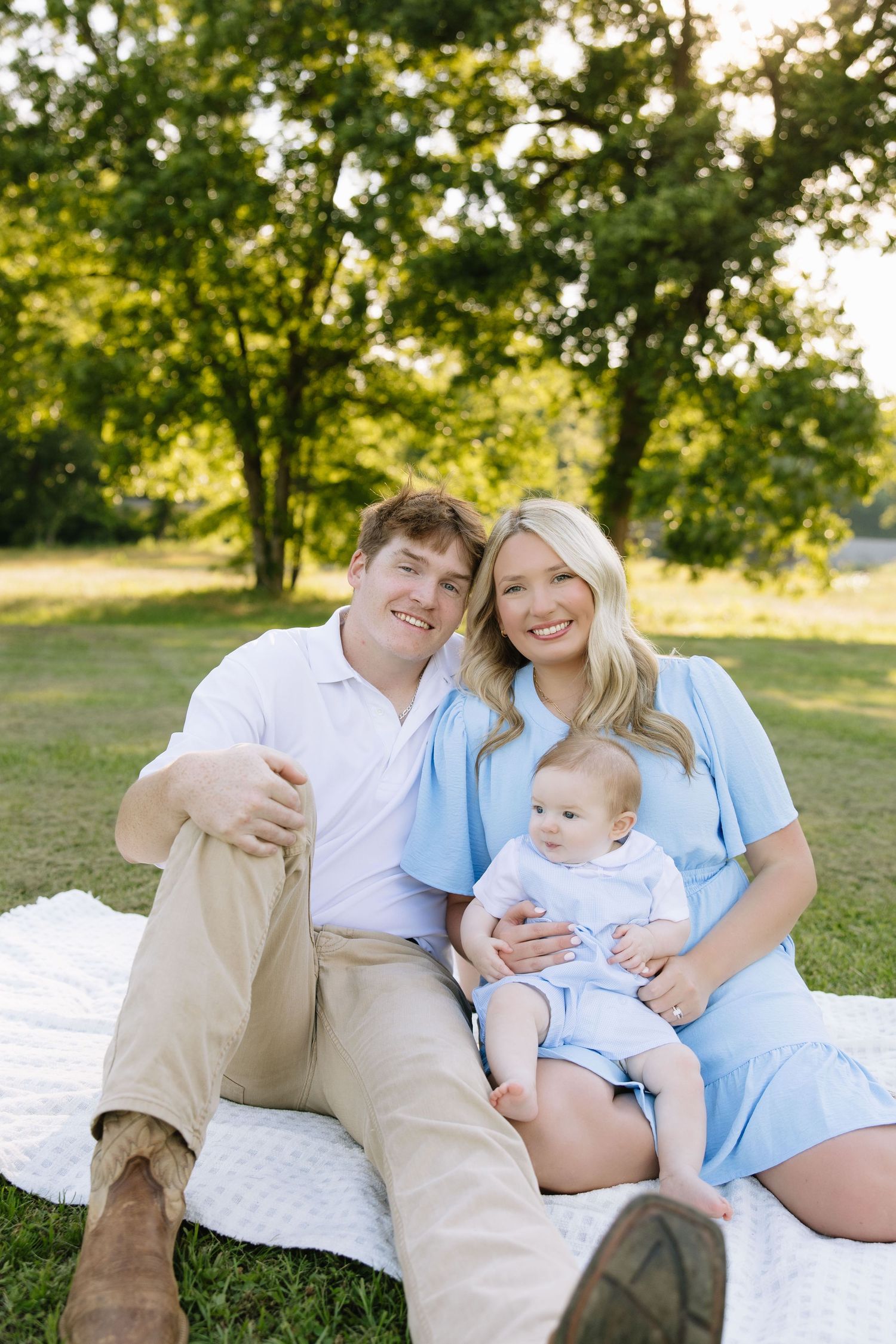 A happy young family sits on a white blanket in a sunny park, with mom and dad smiling while holding their baby.