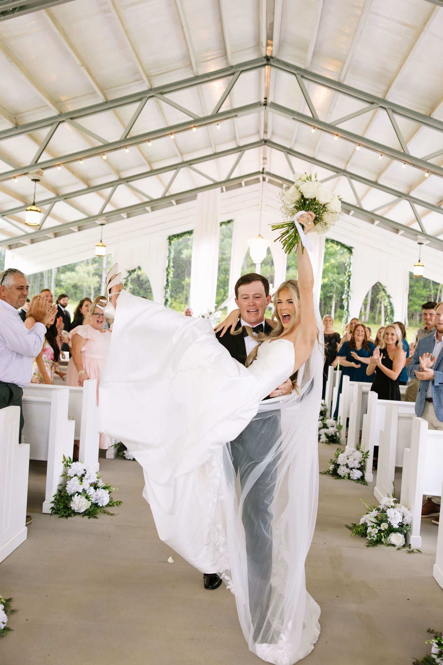Joyful bride and groom celebrate their wedding ceremony exit in a white chapel, as guests cheer and applaud around them.