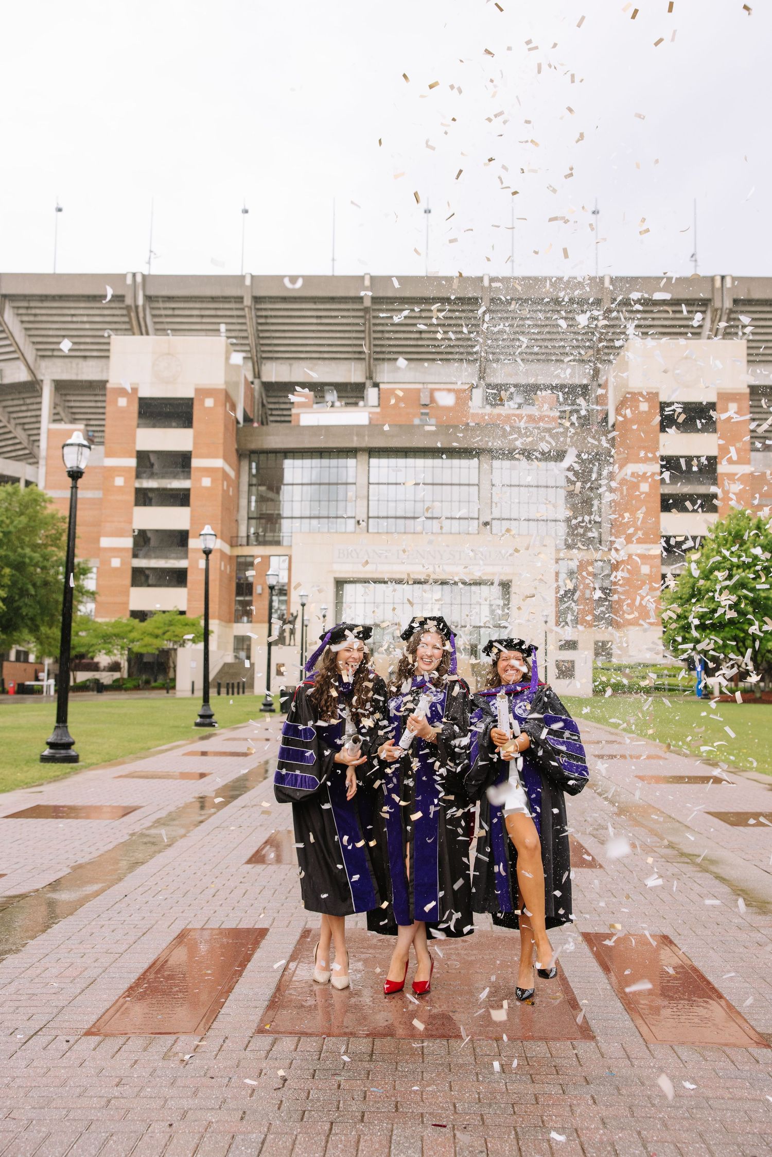Three graduates in purple robes celebrate with confetti outside a university stadium on graduation day.