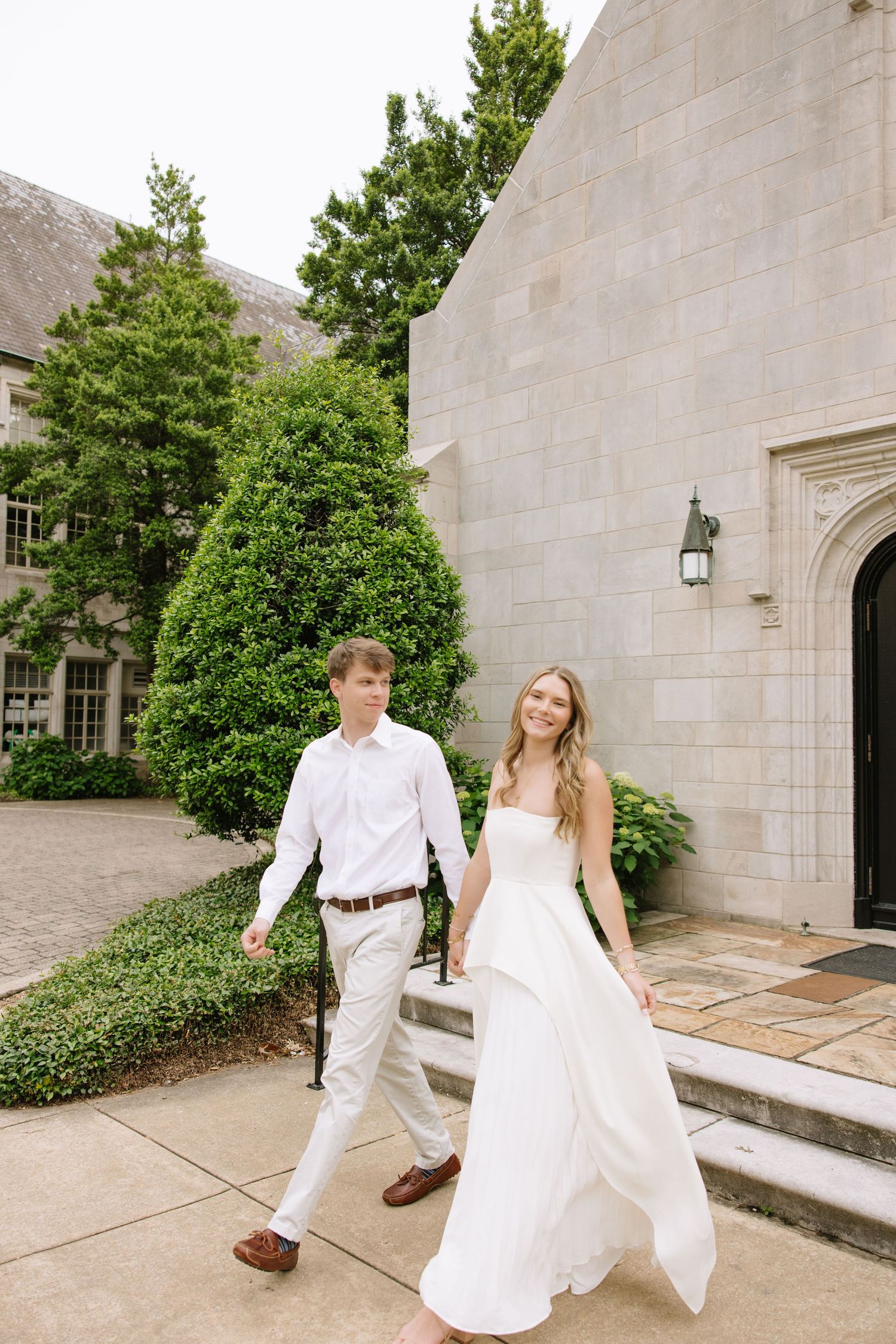 A bride in a white gown and groom in white shirt walk hand-in-hand outside a stone building surrounded by greenery.