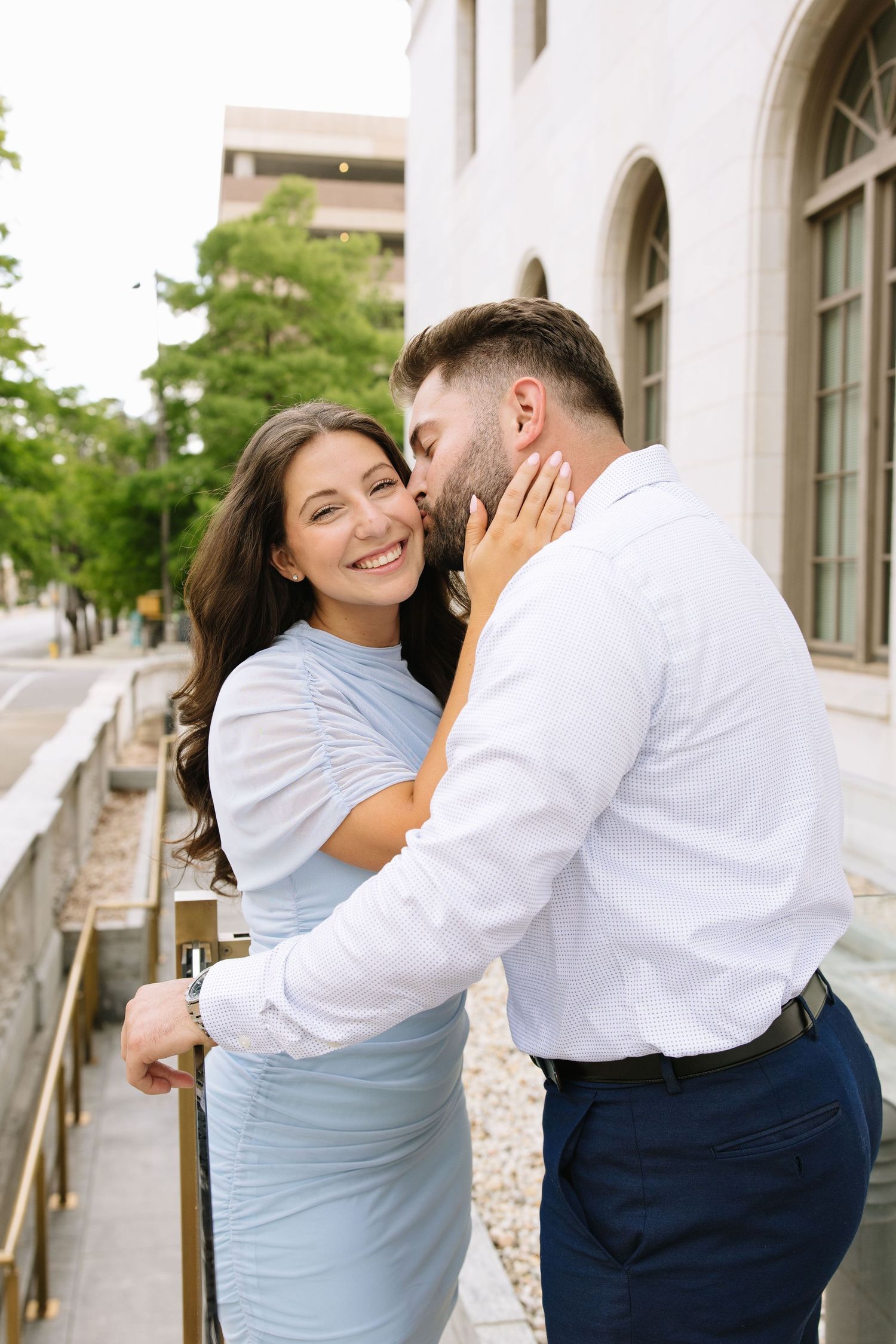 A smiling woman in a light blue dress receives a kiss on the cheek from a man in a white shirt outdoors.