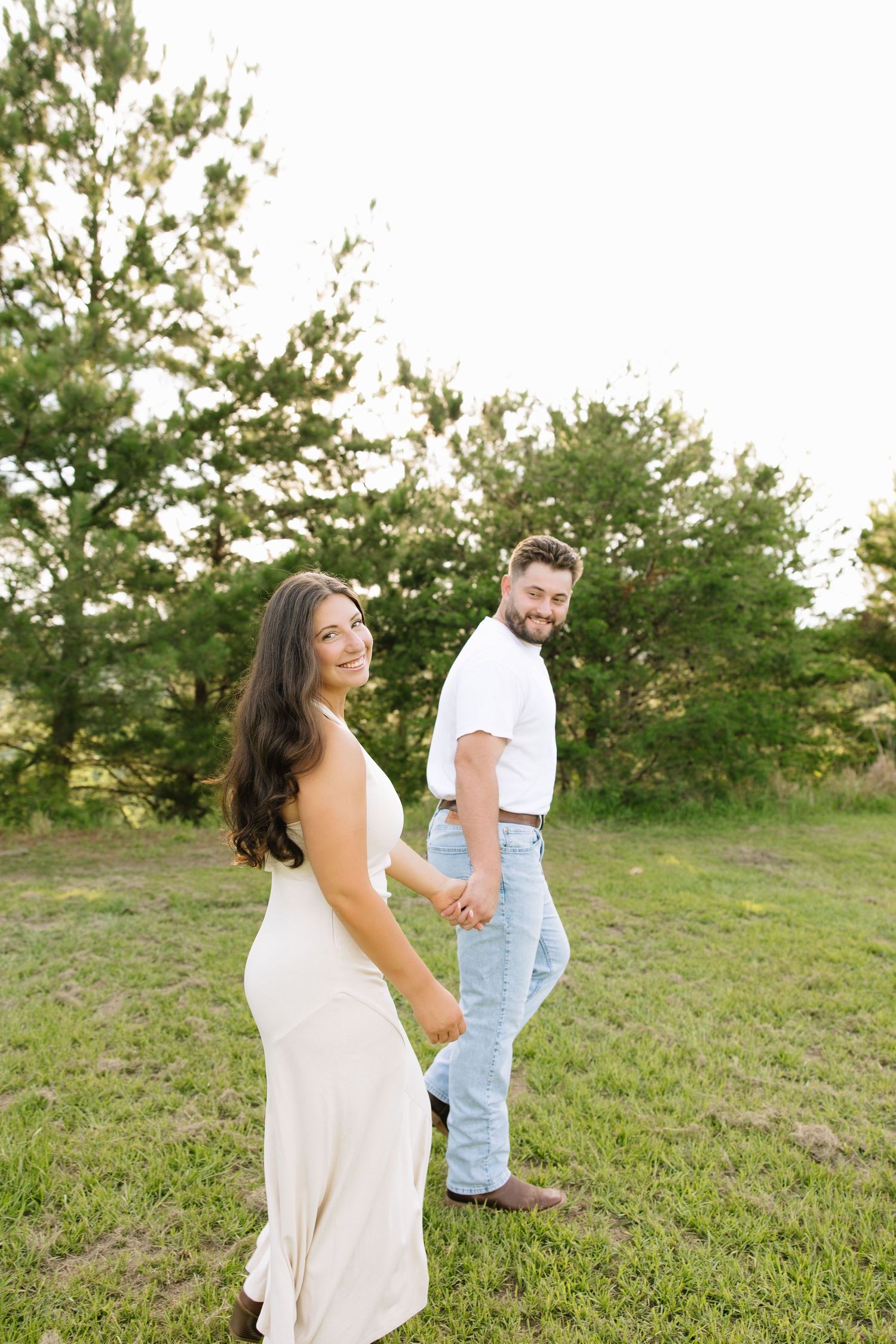 Couple holding hands in a green field, woman in white dress smiling back, man in white shirt and jeans, outdoor engagement photo.