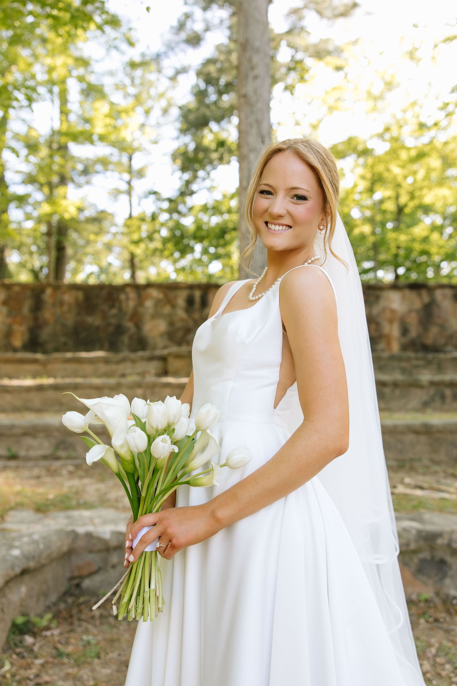 Smiling bride in white gown holding white tulip bouquet with veil outdoors among trees.