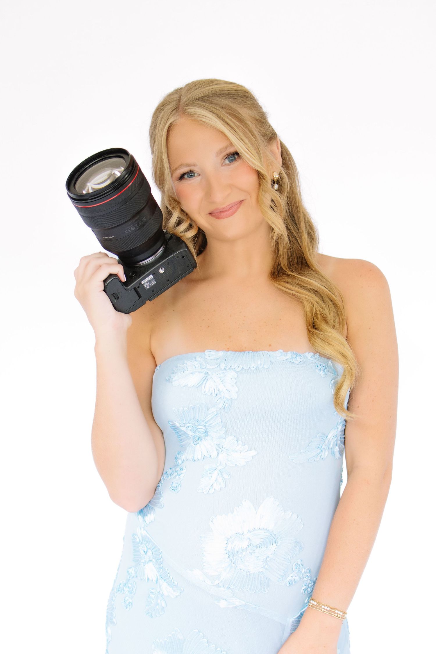 Blonde woman in a light blue strapless floral dress holding a professional Canon DSLR camera with zoom lens.