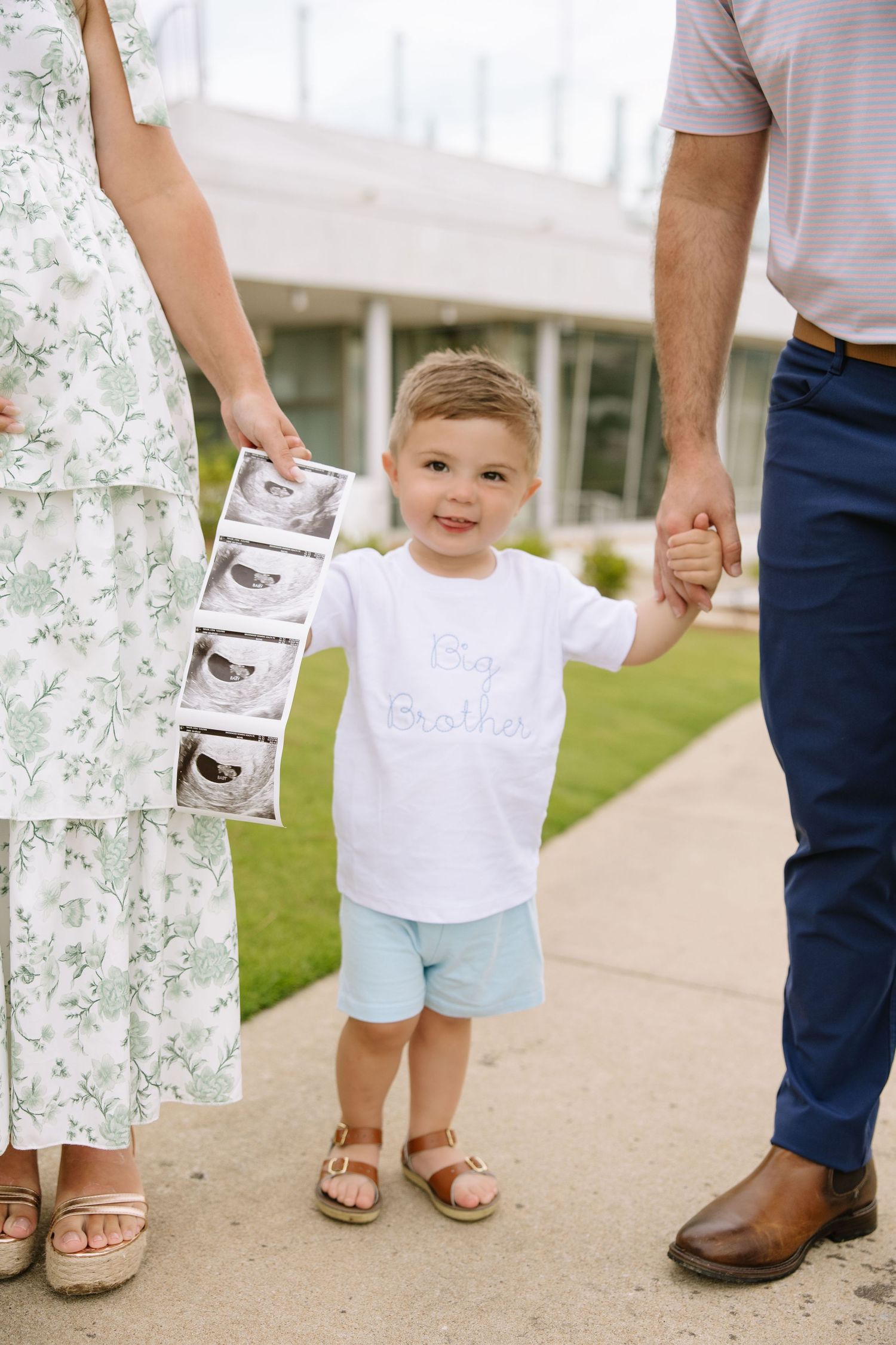 Toddler boy in Big Brother shirt holds parents hands while mom holds ultrasound photos for pregnancy announcement.