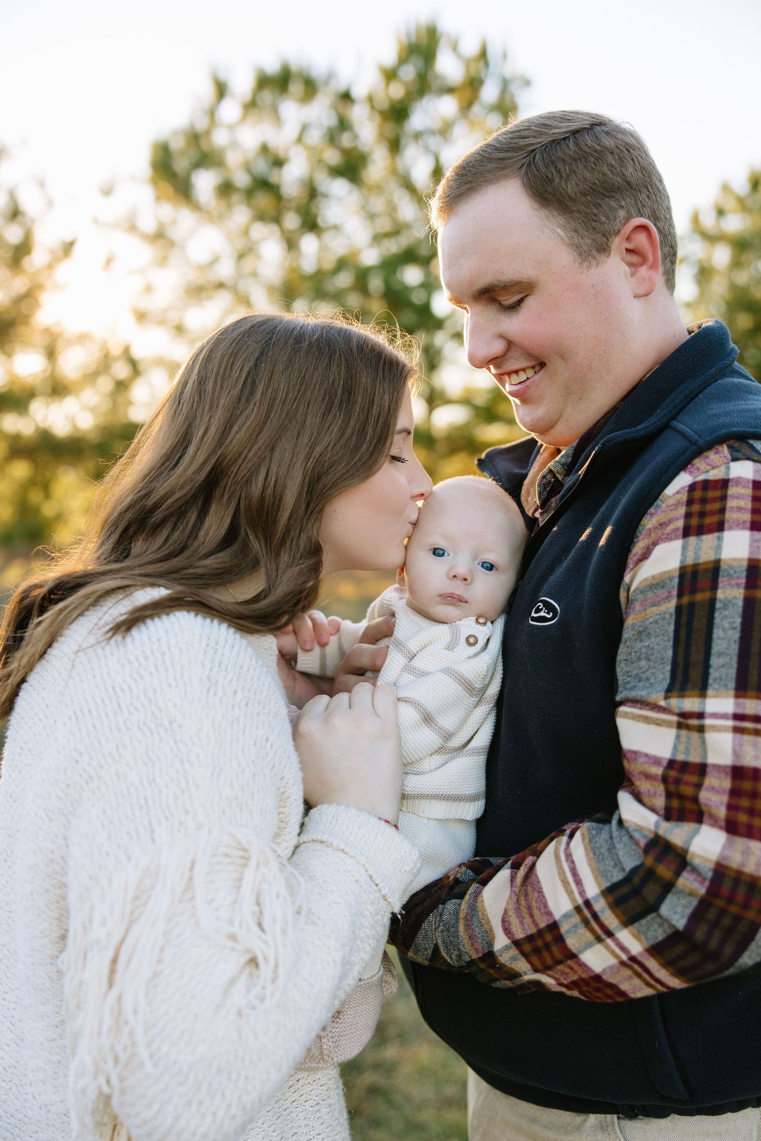 Young parents sharing a tender moment, smiling at their blue-eyed baby outdoors in warm autumn light.