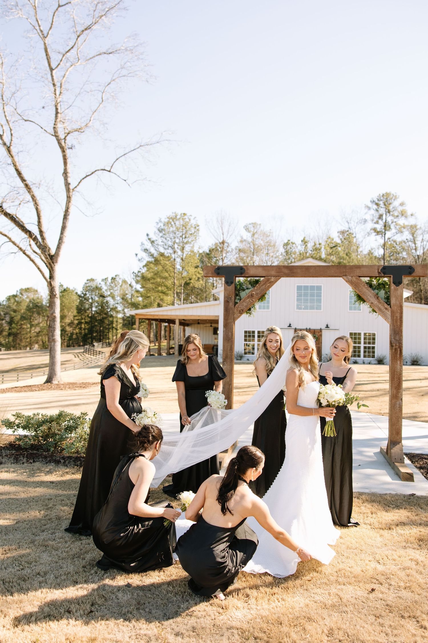 Bridesmaids in black dresses help arrange the bride's gown outdoors at a rustic venue.