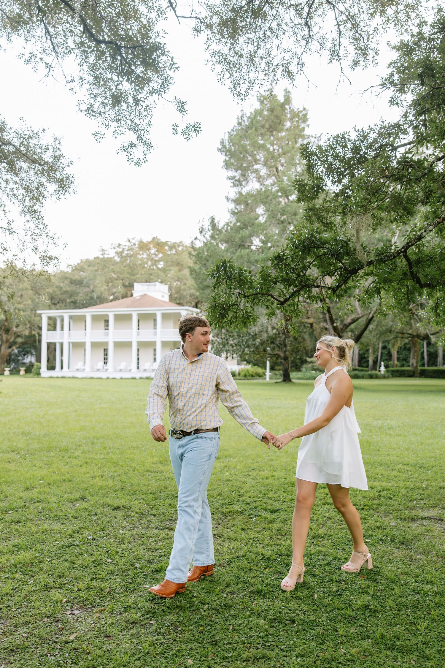A couple holds hands and walks across a lush green lawn in front of a grand white Southern plantation-style home.